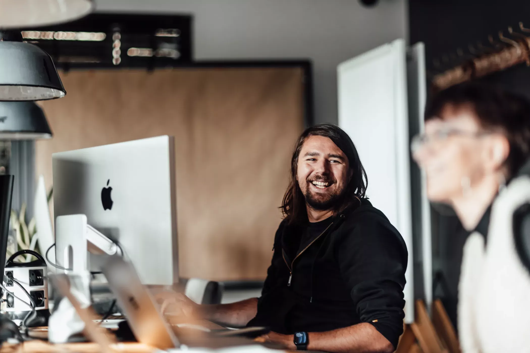 Ein Mitarbeiter der Werbeagentur BICEPS arbeitet an einem iMac im Büro, während eine Kollegin im Vordergrund unscharf zu sehen ist.