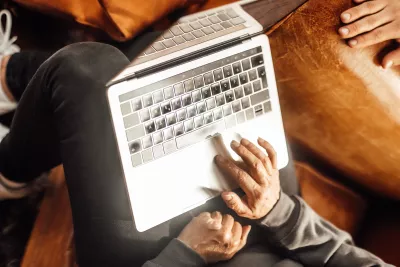 Person sitzt auf einem Sofa und arbeitet mit einem Laptop auf dem Schoß, Hände auf Tastatur und Touchpad, Aufnahme von oben in warmer Umgebung.