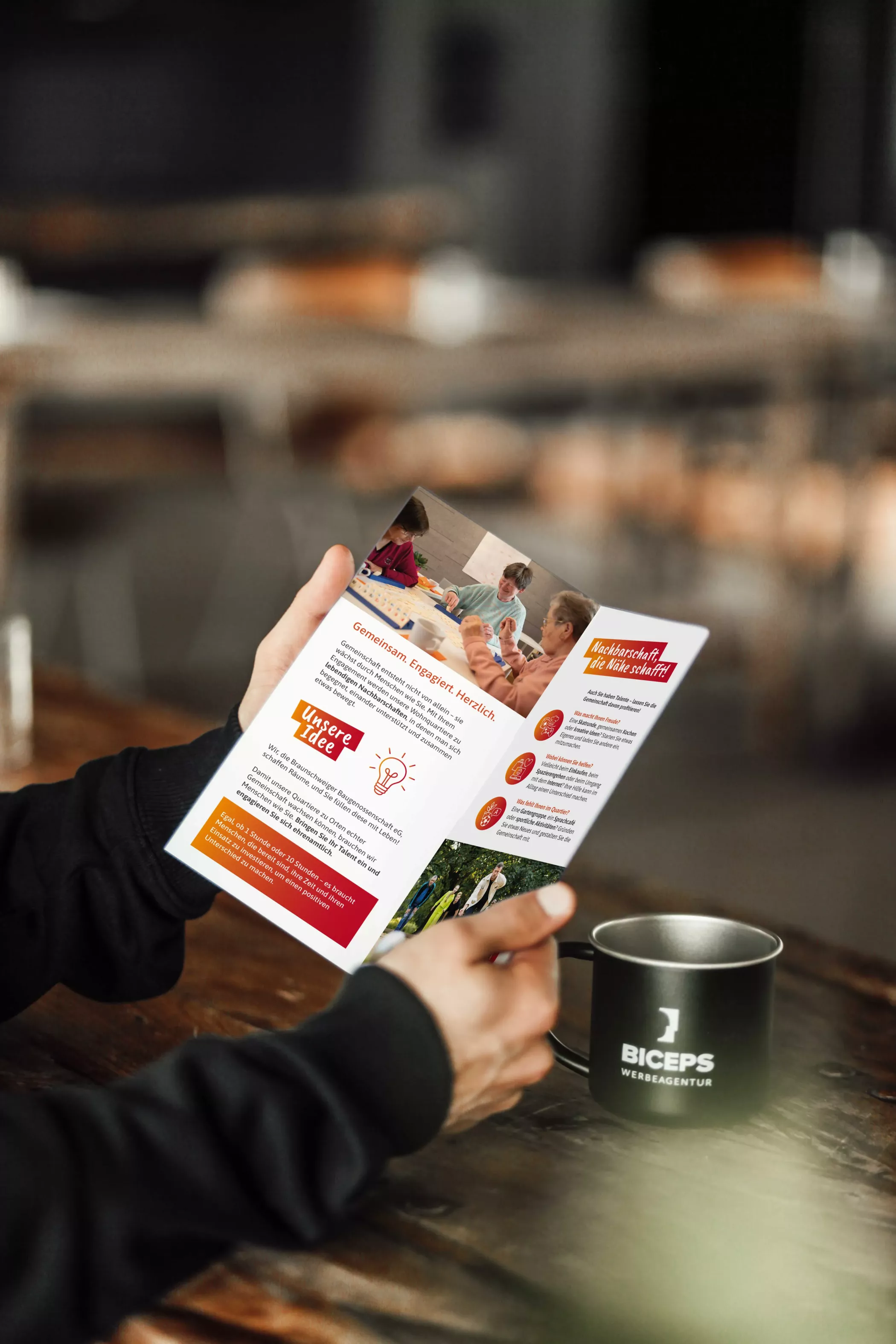 Eine Person hält einen Flyer der Braunschweiger Baugenossenschaft geöffnet in der Hand, während sie an einem Holztisch mit einer schwarzen BICEPS-Tasse sitzt. Auf dem Flyer sind Fotos von Menschen bei gemeinschaftlichen Aktivitäten sowie Texte zur Idee und zum Engagement der Genossenschaft zu sehen.