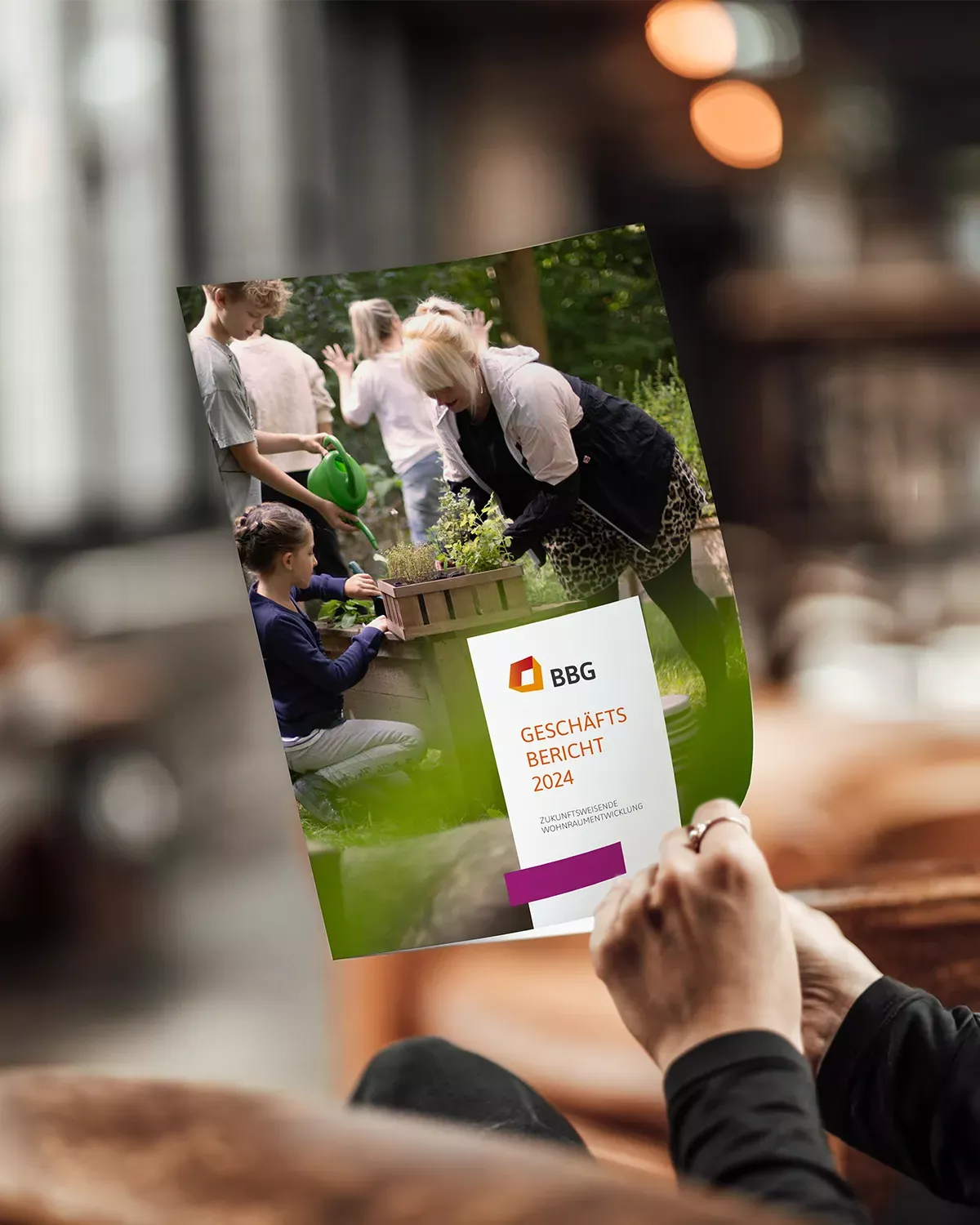 Person hält den Geschäftsbericht 2024 der BBG in der Hand; auf dem Titelbild gärtnern mehrere Menschen gemeinsam in einer grünen Außenanlage.