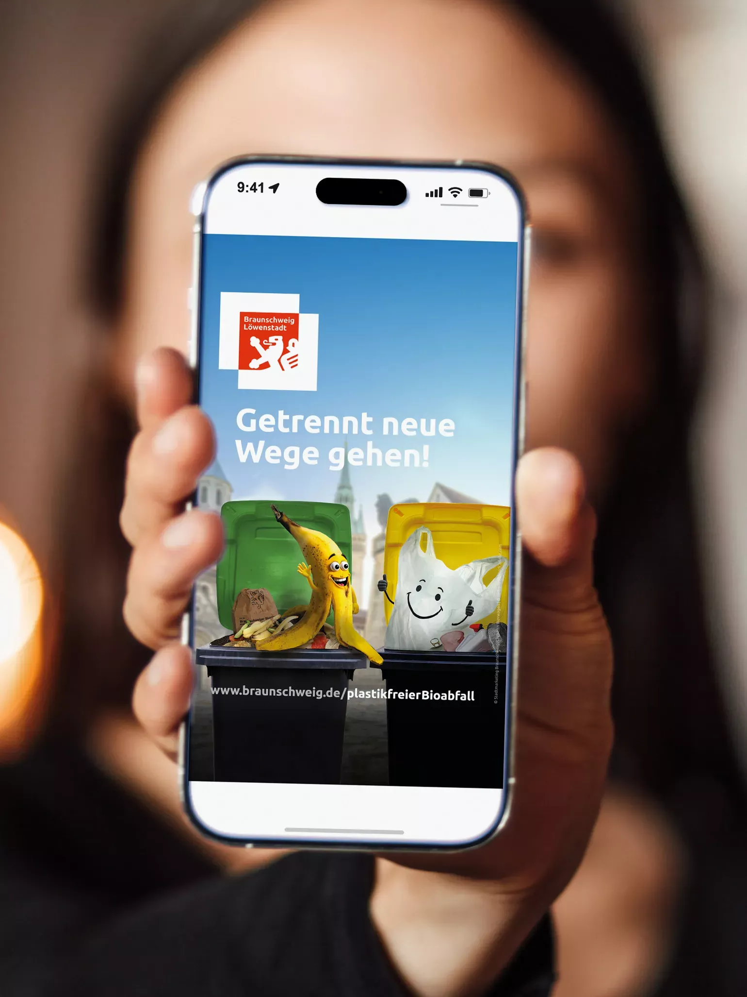 Eine Person hält Smartphone mit geöffneter Kampagnenseite der Stadt Braunschweig zur Biomüllkampagne ‚Getrennt neue Wege gehen!‘. Auf dem Screen ist das Kampagnenmotiv mit Biotonne, Gelber Sack, animierter Bananenschale und Slogan vor Stadtsilhouette zu sehen.