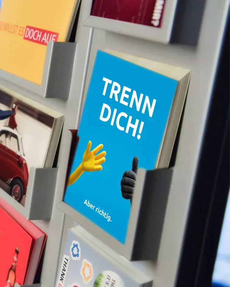 Citycard mit der Aufschrift „TRENN DICH! Aber richtig.“ in einem Postkartenständer. Die Karte zeigt eine gelbe und eine schwarze Hand als Symbol für richtige Mülltrennung. Die Kampagnenkarte steht zwischen anderen bunten Postkarten und ist prominent platziert.