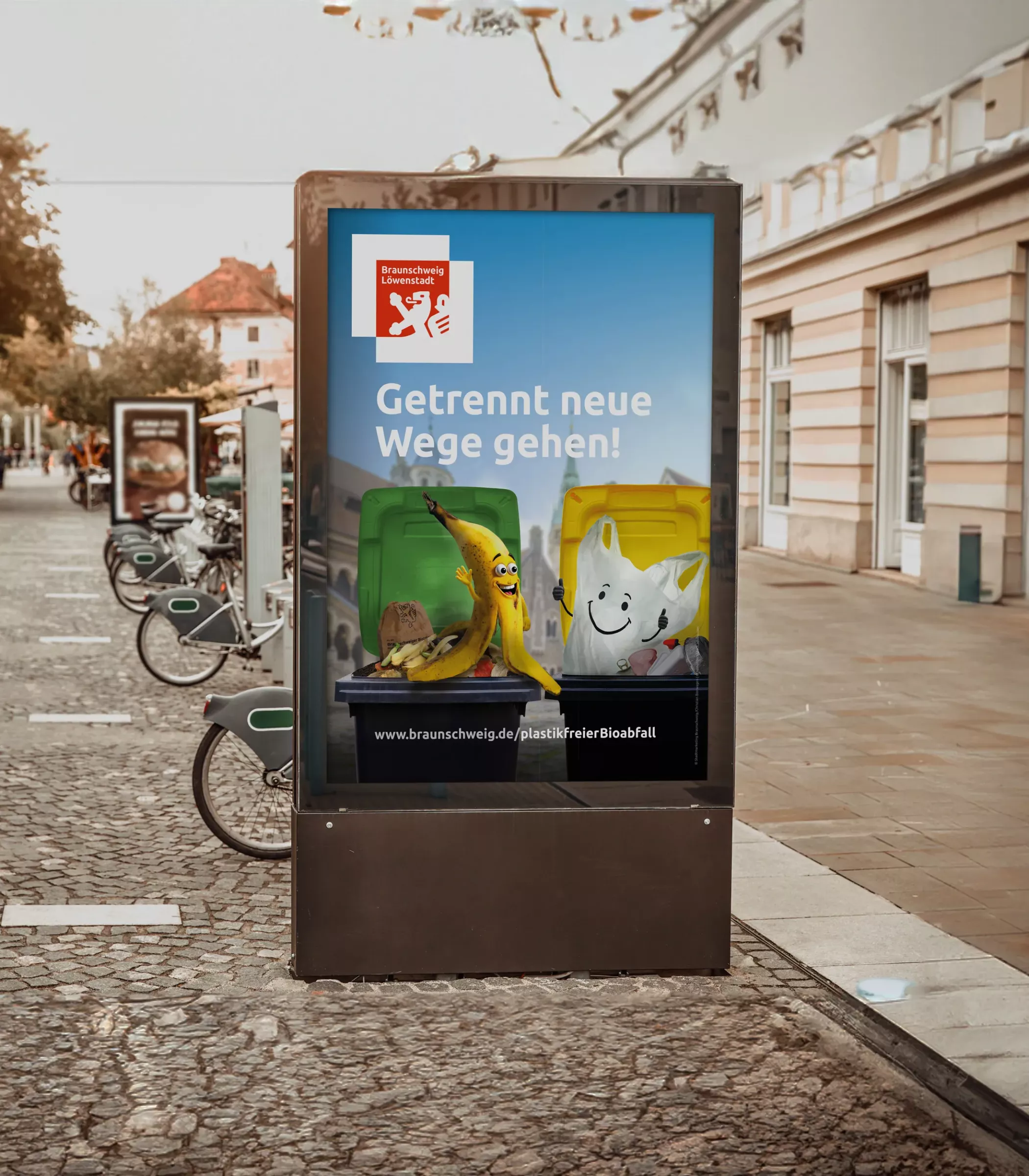 Digitale Citylight-Werbefläche in einer belebten Fußgängerzone von Braunschweig mit dem Kampagnenmotiv der Biomüll-Kampagne „Getrennt neue Wege gehen!“. Auf dem Plakat zu sehen: eine gelbe Biotonne mit einer animierten Bananenschale und eine gelbe Tonne mit einer animierten Plastiktüte vor städtischer Kulisse.