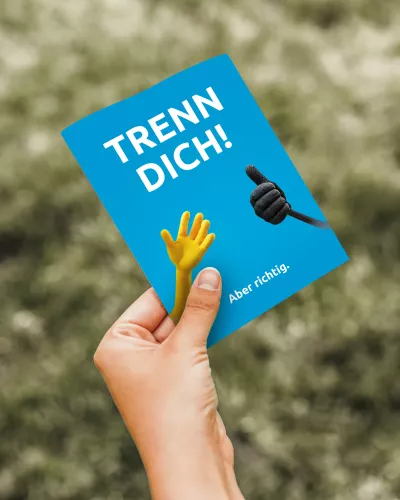 Eine Person hält eine Citycard der Biomüllkampagne in der Hand. Die Karte zeigt die Botschaft „TRENN DICH! Aber richtig.“ auf blauem Hintergrund. Zwei stilisierte Hände – eine gelb, eine schwarz – symbolisieren die Trennung von Bio- und Restmüll.