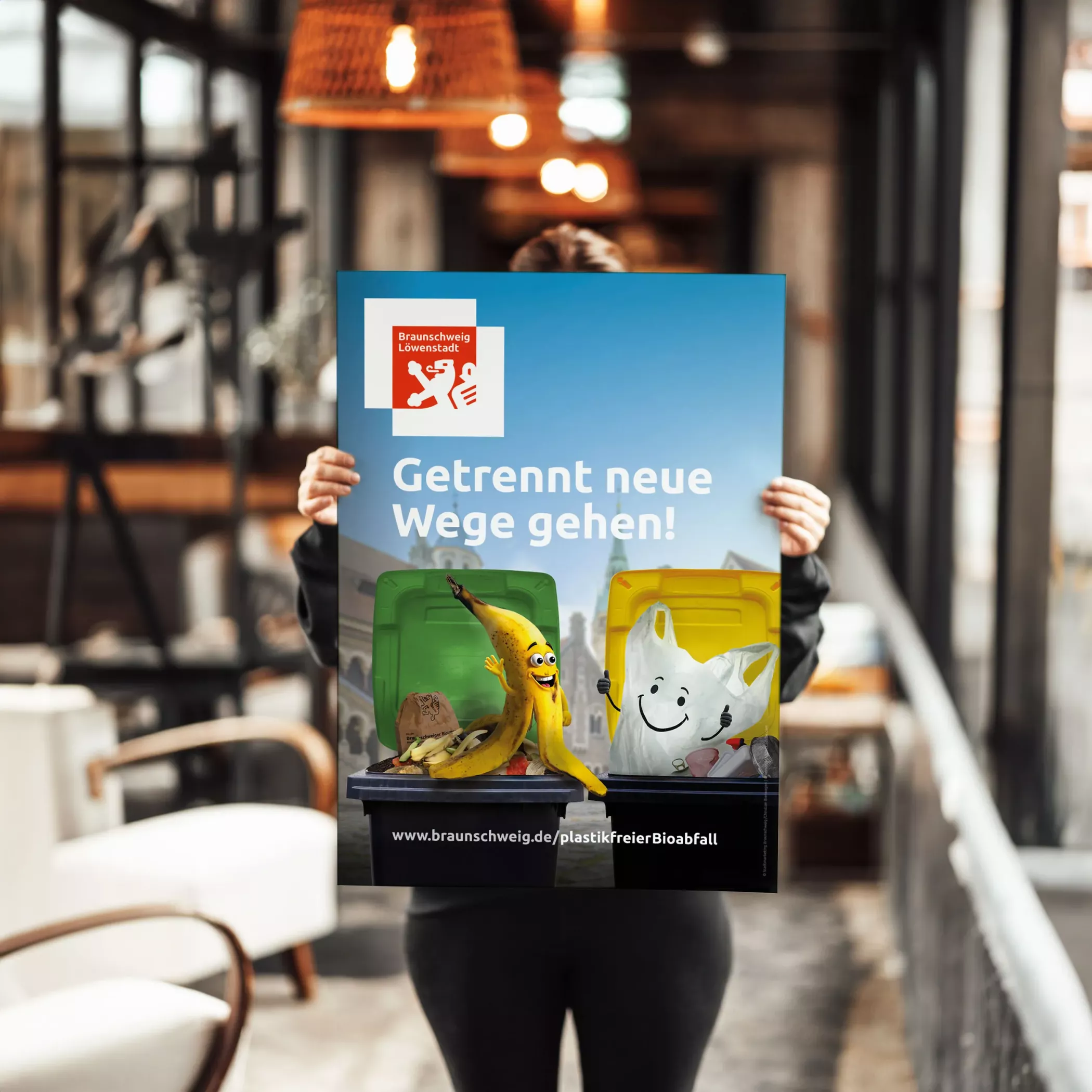 Eine Person hält eine Großflächenplakat-Version der Biomüllkampagne der Stadt Braunschweig in den Händen. Auf dem Plakat sind eine animierte Bananenschale und eine Plastiktüte zu sehen, die jeweils fröhlich in Richtung ihrer passenden Mülltonne winken. Darüber der Slogan „Getrennt neue Wege gehen!“.