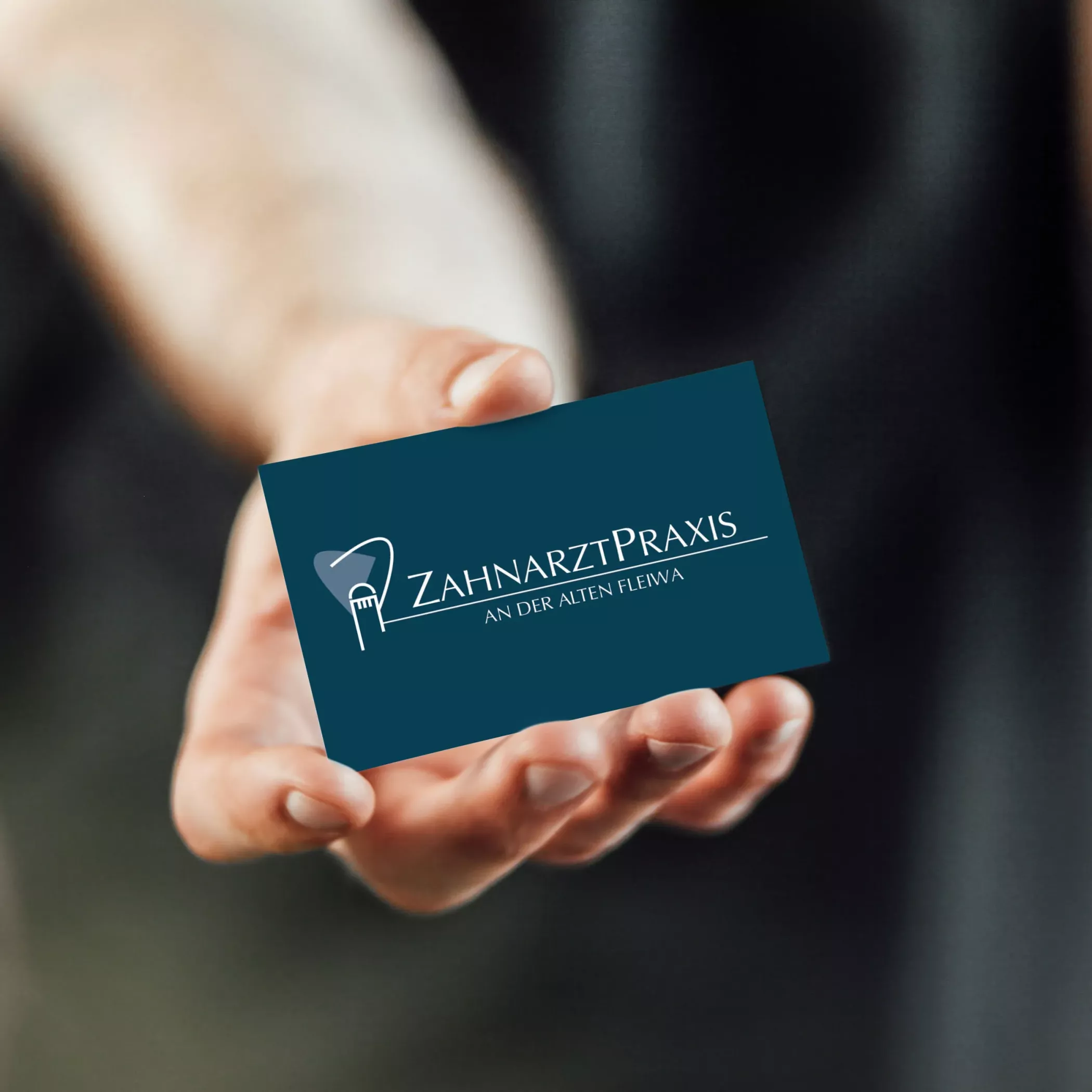 Eine Hand hält eine dunkelblaue Visitenkarte der „Zahnarztpraxis an der Alten Fleiwa“. Auf der Karte ist das Logo mit stilisierter Darstellung der Alten Fleiwa und einer Zahnbürste integriert, dazu der Schriftzug der Praxis in weißer Schrift. Das Design wirkt modern, hochwertig und lokal verankert.