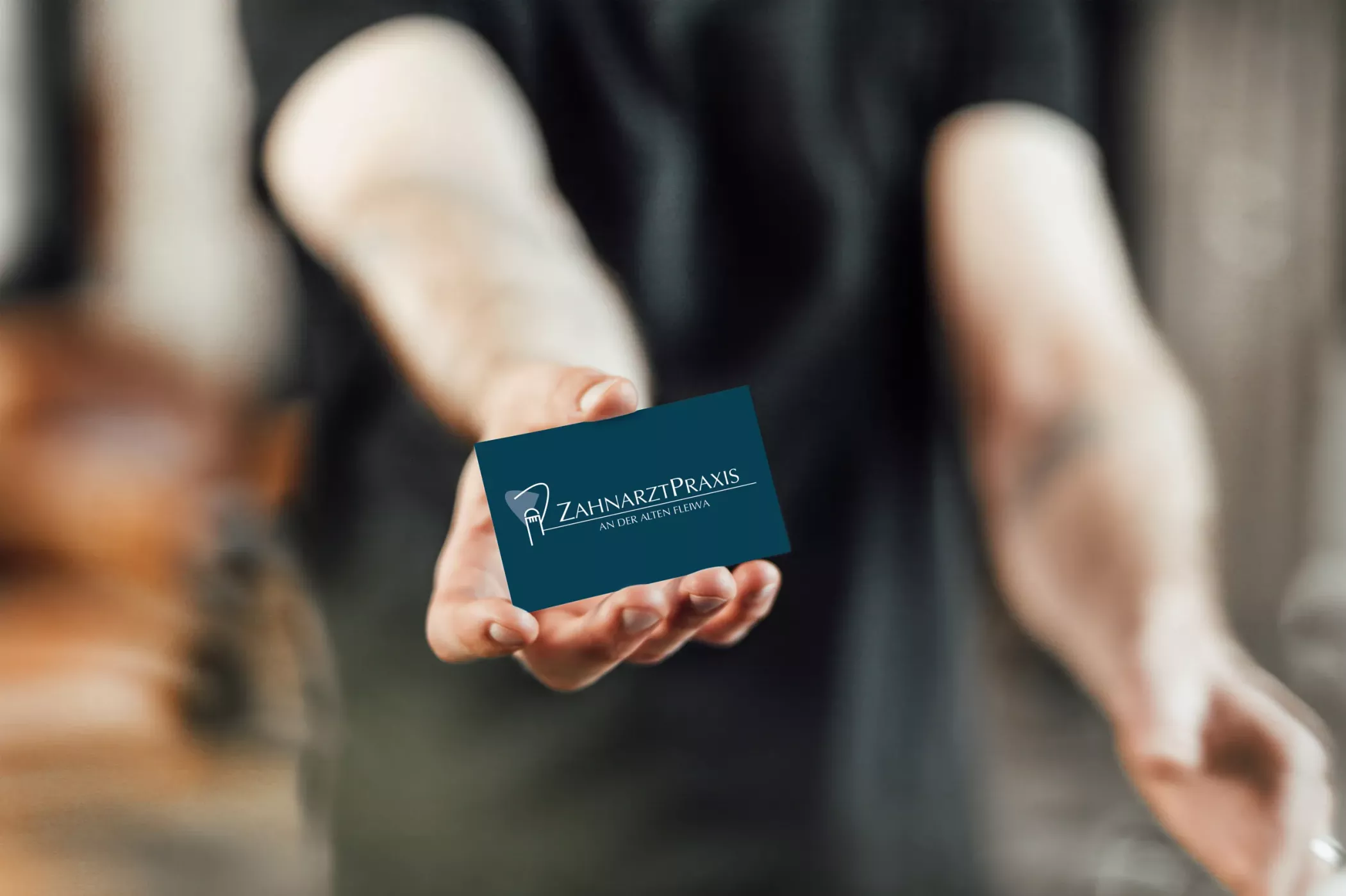 Eine Person hält eine dunkelblaue Visitenkarte der „Zahnarztpraxis an der Alten Fleiwa“ in beiden Händen. Auf der Karte ist das Praxislogo mit der Silhouette der Alten Fleiwa in Hannover sowie der Schriftzug der Praxis zu sehen. Das Design wirkt modern, professionell und lokal verankert.