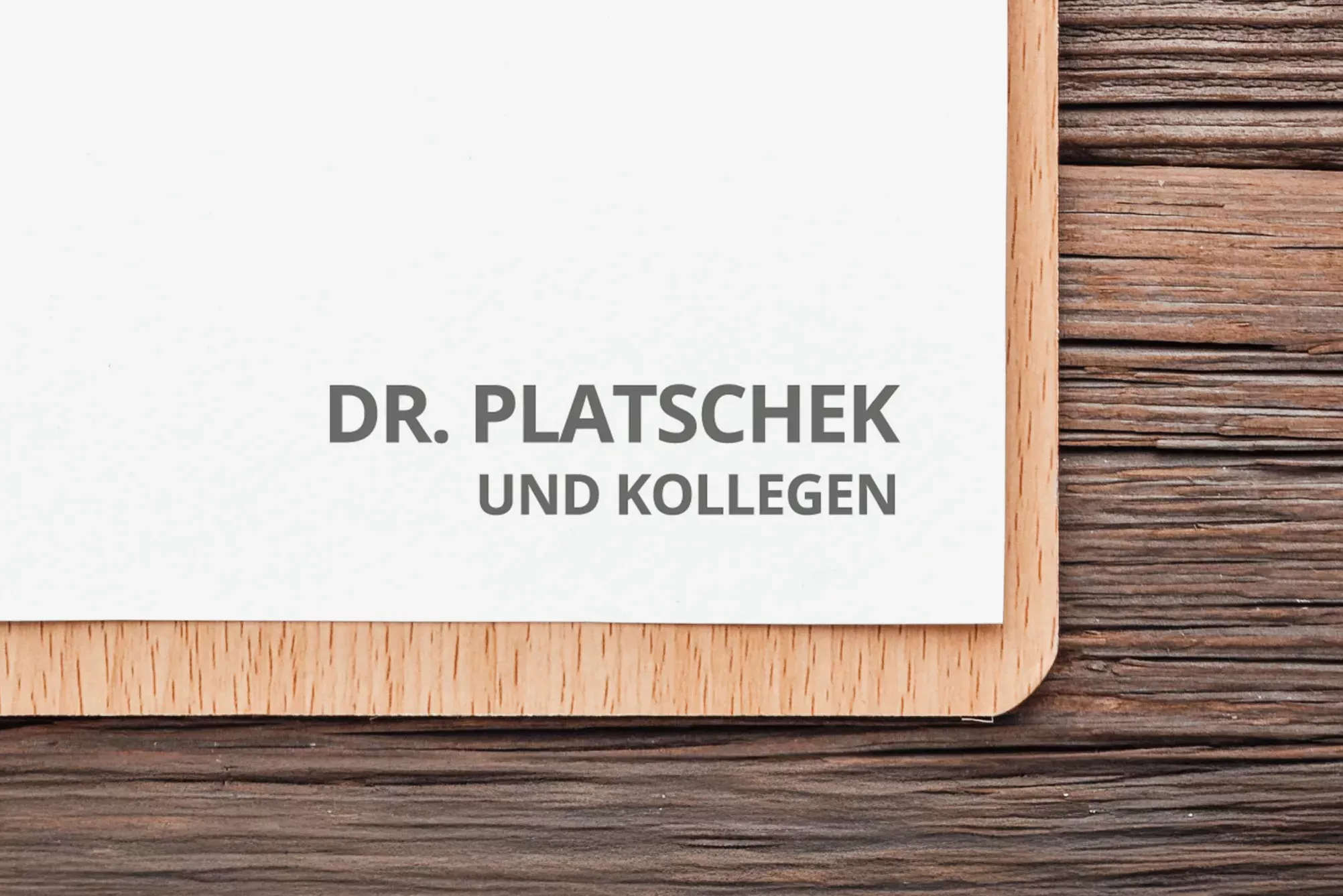 Ausschnitt eines Klemmbretts auf einer Holzoberfläche mit einem Papierbogen, auf dem das Logo „Dr. Platschek und Kollegen“ in grauer Schrift abgebildet ist. Das Design wirkt minimalistisch, klar und professionell.