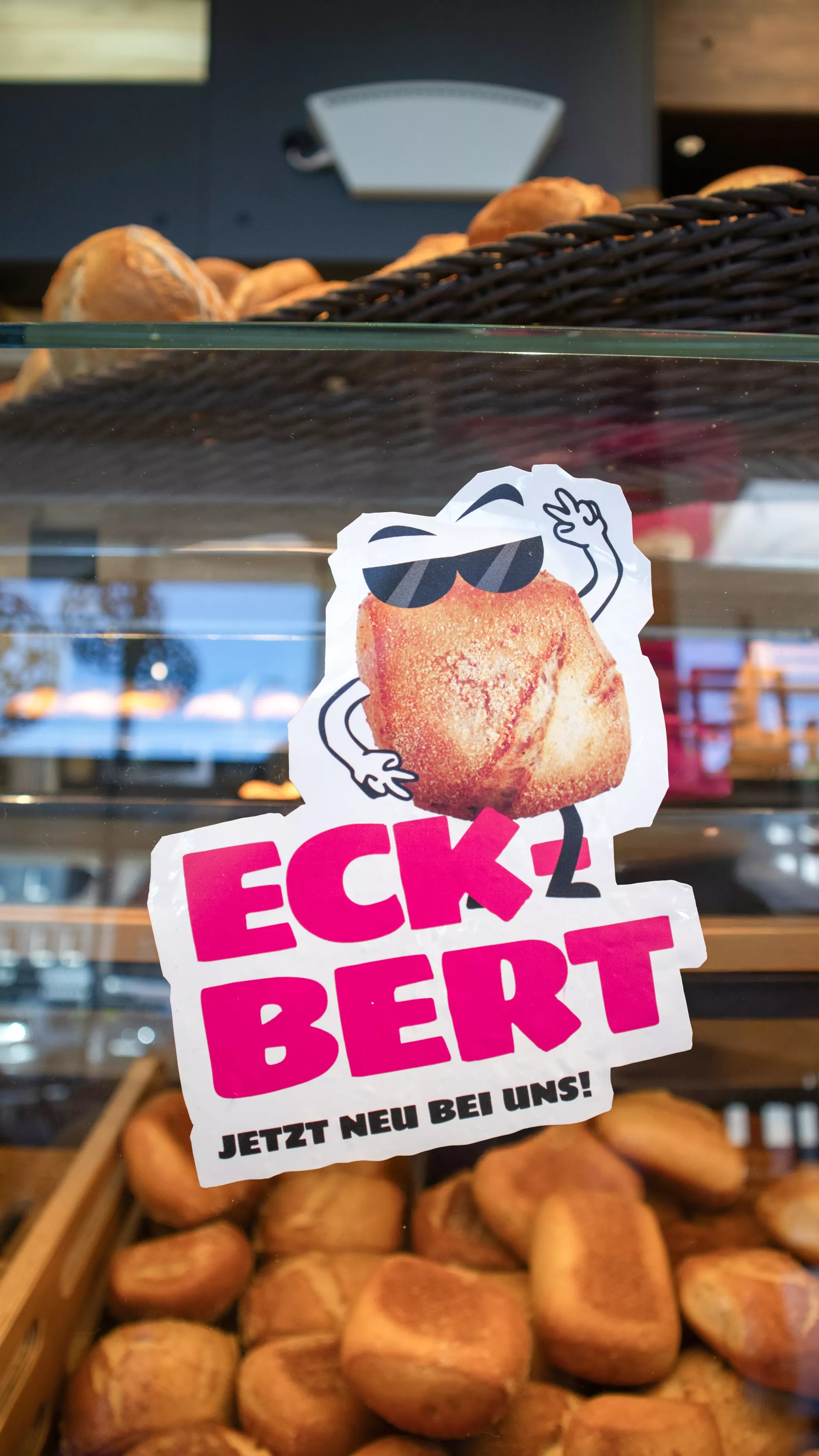 Aufkleber mit Brötchen-Comicfigur „Eckbert“ an der Glasscheibe einer Bäckereitheke. Im Hintergrund sind Brötchen in der Auslage zu sehen. Der Aufkleber zeigt Eckbert mit Sonnenbrille und erhobener Hand, darunter steht: „Eckbert – jetzt neu bei uns!“.