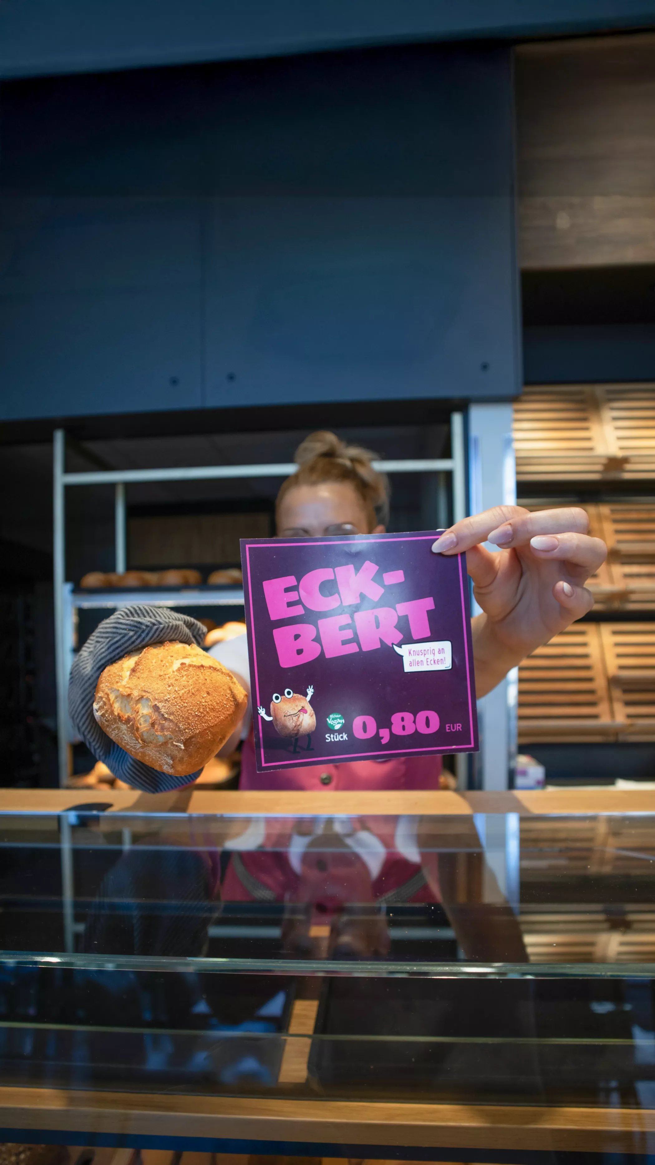 Eine Verkäuferin hält ein Preisschild für das Brötchen „Eckbert“ in die Kamera. Auf dem Schild ist der Preis von 0,80 € zu sehen sowie ein gezeichneter Eckbert mit Text: „Knusprig in eine Linie“. In der anderen Hand hält sie das passende Brötchen.