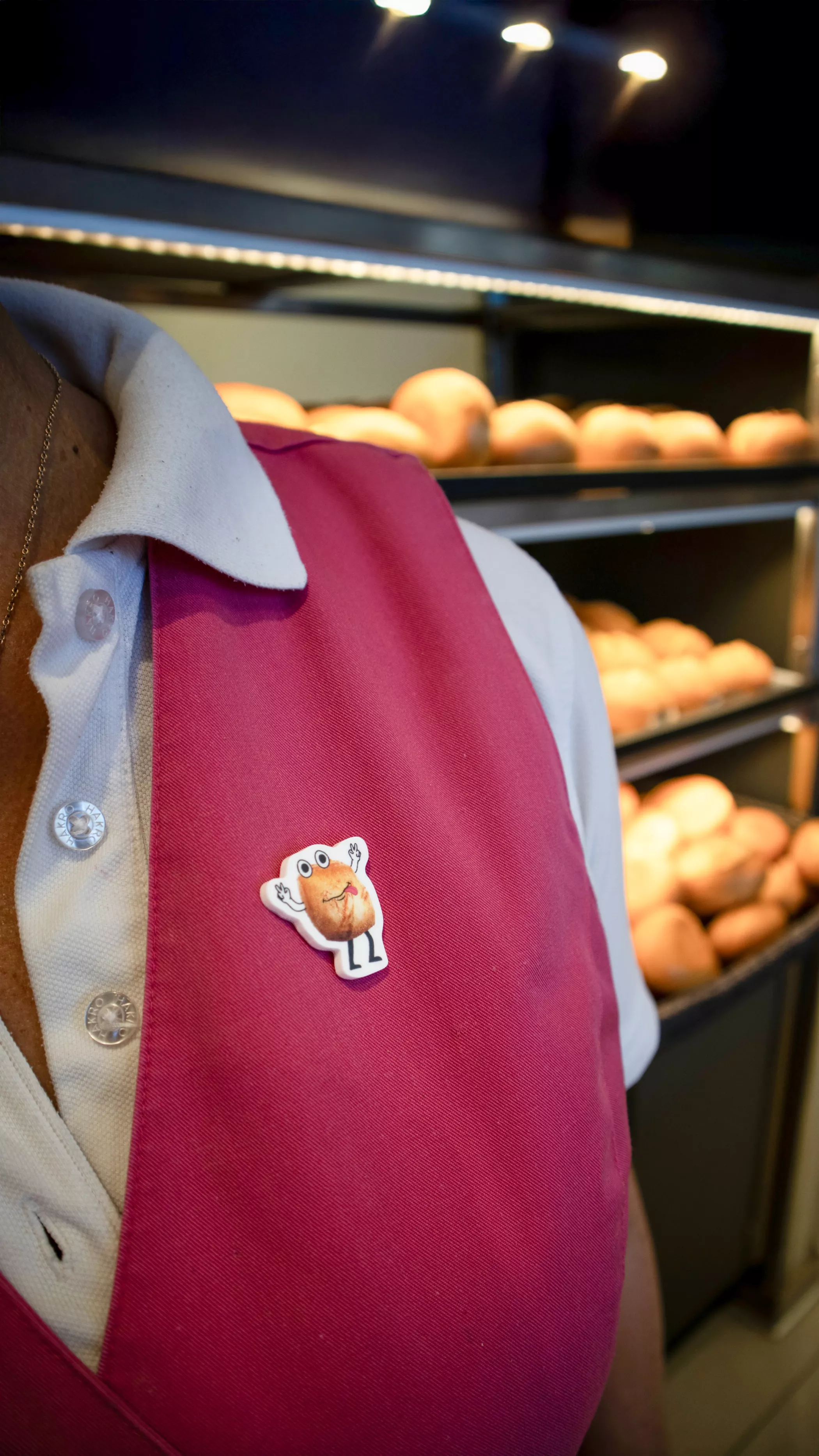 Nahaufnahme einer Mitarbeiterin der Altstadtbäckerei Richter in Uniform, auf deren Schürze ein Button mit dem Comic-Maskottchen „Eckbert“ befestigt ist; im Hintergrund sind Brote im Verkaufsregal zu sehen.