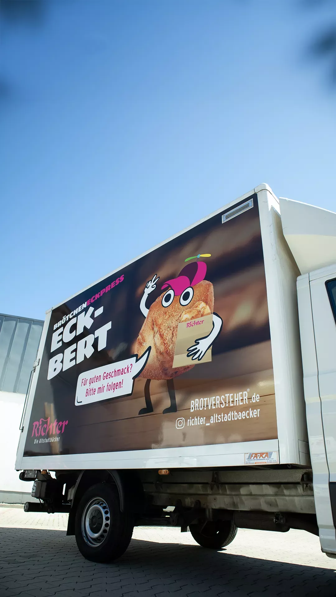 Ein weißer Lieferwagen der Altstadtbäckerei Richter mit großflächiger Werbegrafik für das Brötchen „Eckbert“. Das Brötchen ist als sympathische Comicfigur mit Armen, Beinen, Augen und pinker Tolle dargestellt und hält eine Brottüte mit Richter-Logo. Daneben steht der Schriftzug „Brötcheneckpress Eckbert“ und eine Sprechblase mit dem Text: „Für guten Geschmack? Bitte mir folgen!“ Auf dem unteren Bereich befinden sich Website-URL und Instagram-Handle.