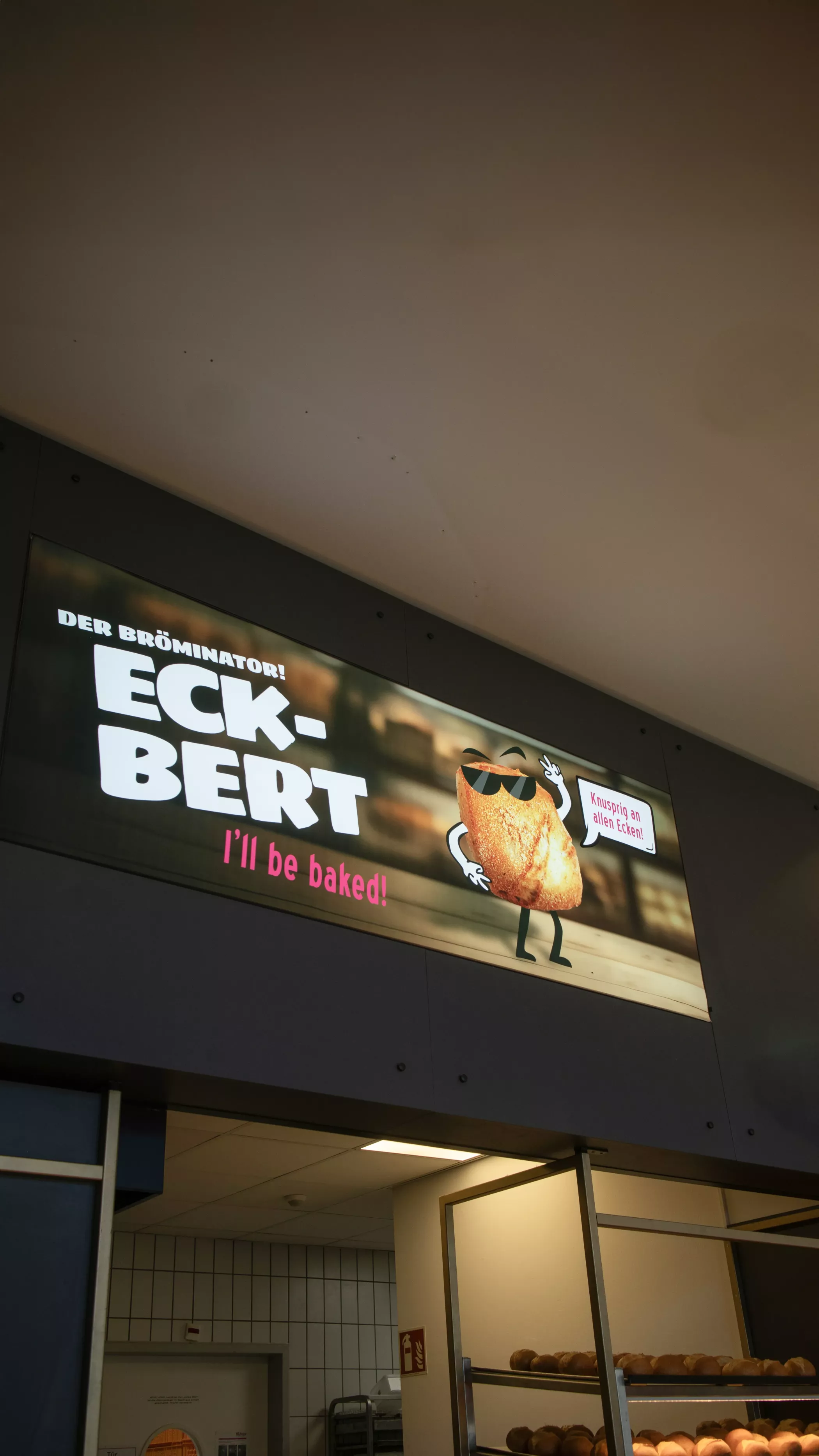 In der Filiale der Altstadtbäckerei Richter zeigt ein beleuchteter Leuchtrahmen das Eckbert-Maskottchen mit Sonnenbrille und dem Spruch „I’ll be baked!“. Darüber steht „Der Bröminator!”, daneben befindet sich ein Schild mit der Aufschrift „Knusprig ist keine Option”. Unten ist der Tresenbereich der Filiale zu sehen.