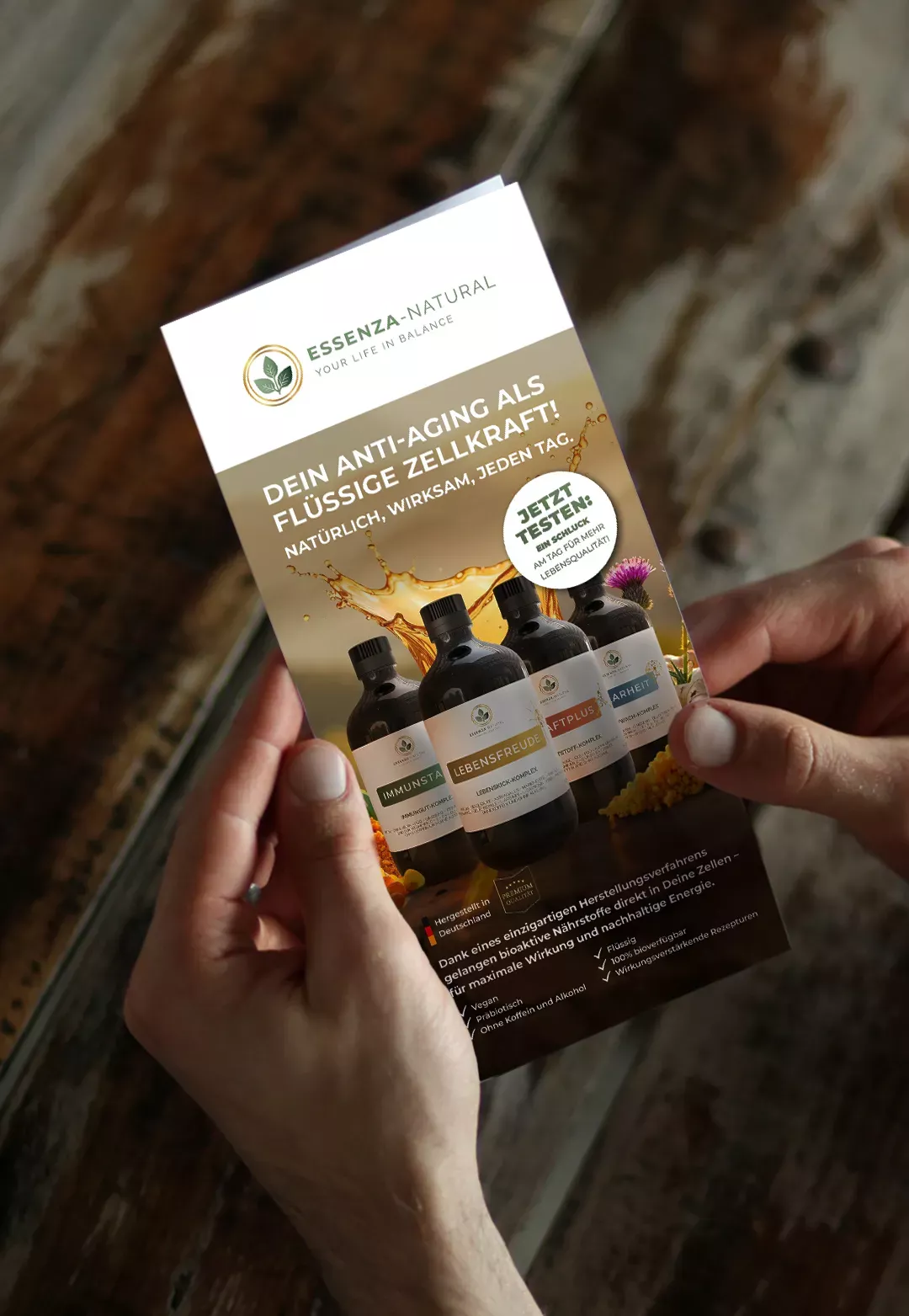 Person hält einen Essenza-Natural-Flyer mit Anti-Aging-Produktmotiv und Informationen zu flüssigen Nahrungsergänzungsmitteln.