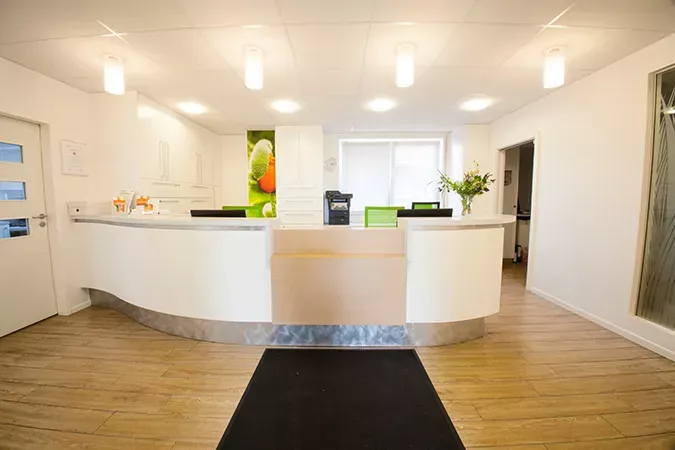 Moderner Empfangsbereich der Frauenarztpraxis Dr. Altevogt und Rühmkorb-Claassen mit geschwungenem weißen Tresen, grünen Akzentstühlen und heller Holzoptik. Im Hintergrund sorgen florale Wandbilder und eine freundliche Beleuchtung für eine einladende Atmosphäre.