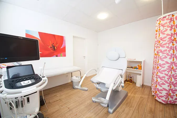 Behandlungsraum der Frauenarztpraxis Dr. Altevogt und Rühmkorb-Claassen mit moderner Untersuchungseinheit, Ultraschallgerät und Liege. Der Raum ist hell gestaltet, mit Holzoptik-Boden, weißen Wänden, einem roten Blumenbild und einer gemusterten Sichtschutzwand. Die Atmosphäre wirkt freundlich, professionell und einladend.