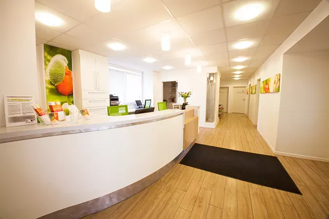 Empfangsbereich der Frauenarztpraxis Dr. Altevogt und Rühmkorb-Claassen mit modernem weißen Tresen, grünen Akzenten und freundlicher Beleuchtung. An den Wänden hängen farbenfrohe Blumenbilder, der Boden ist in heller Holzoptik gestaltet. Die Atmosphäre wirkt einladend, hell und professionell.