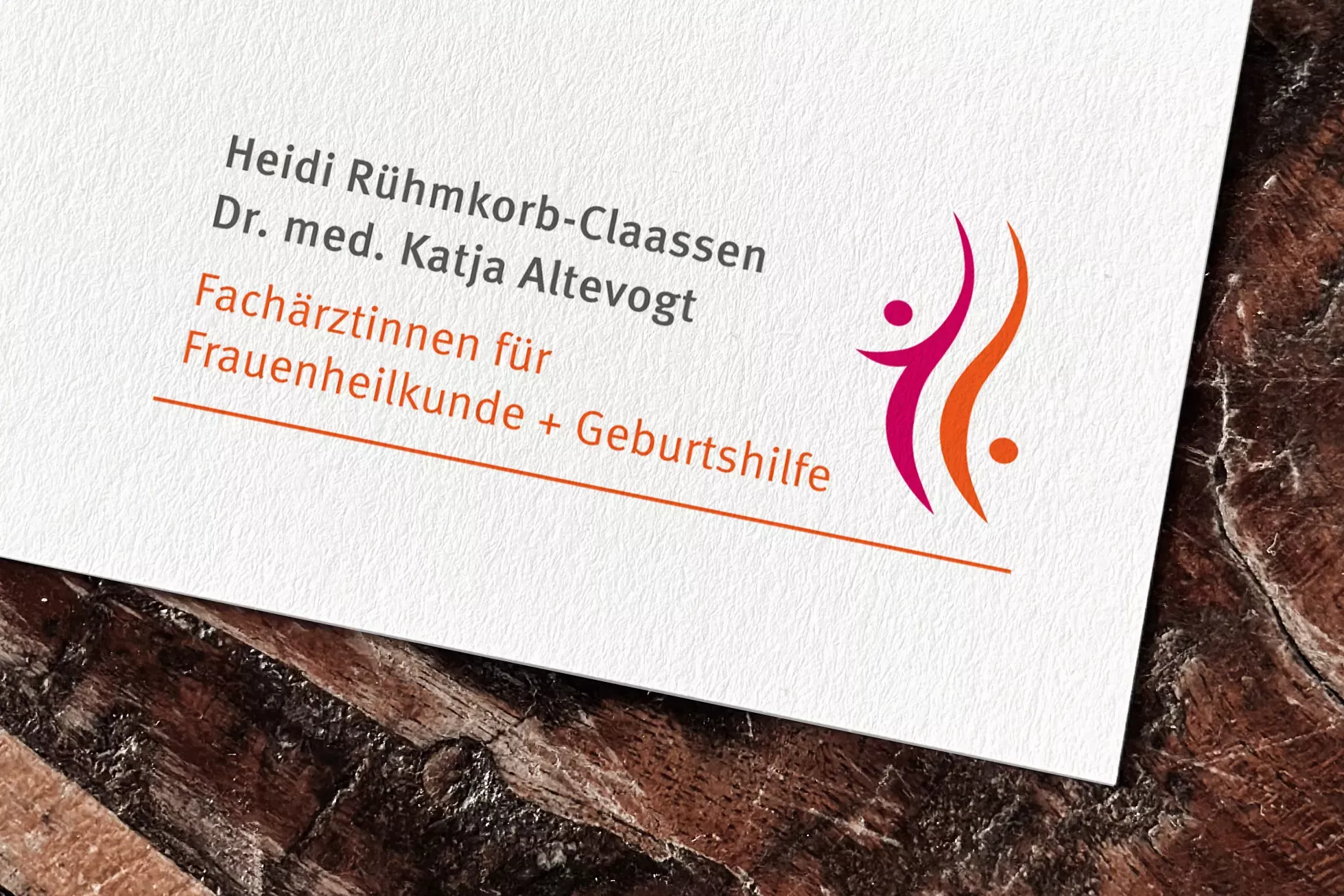 Nahaufnahme eines Druckdokuments der Frauenarztpraxis Heidi Rühmkorb-Claassen und Dr. med. Katja Altevogt. Zu sehen sind die Namen der Ärztinnen, die Berufsbezeichnung „Fachärztinnen für Frauenheilkunde + Geburtshilfe“ in oranger Schrift sowie ein stilisiertes Logo mit zwei abstrakten Figuren in Magenta und Orange auf weißem Hintergrund. Das Design wirkt modern, freundlich und vertrauensvoll.