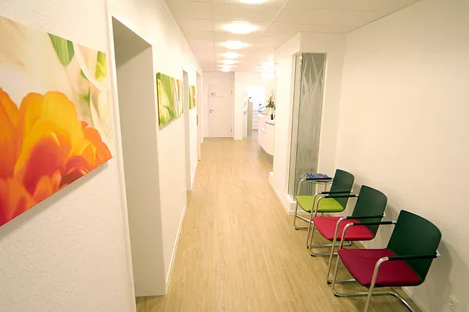 Flur der Frauenarztpraxis Dr. Altevogt und Rühmkorb-Claassen mit heller Holzoptik, weißen Wänden und farbenfrohen Blumenbildern. Rechts stehen moderne Stühle in Grün und Rot, die auf das Farbkonzept der Praxis abgestimmt sind. Der Flur wirkt freundlich, hell und einladend.