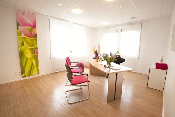 Helles Sprechzimmer der Frauenarztpraxis Dr. Altevogt und Rühmkorb-Claassen mit großen Fenstern, einem modernen Schreibtisch und Stühlen mit pinkfarbenen Polstern. An der Wand hängt ein großes florales Kunstwerk, das die freundliche und beruhigende Atmosphäre des Raumes unterstreicht.