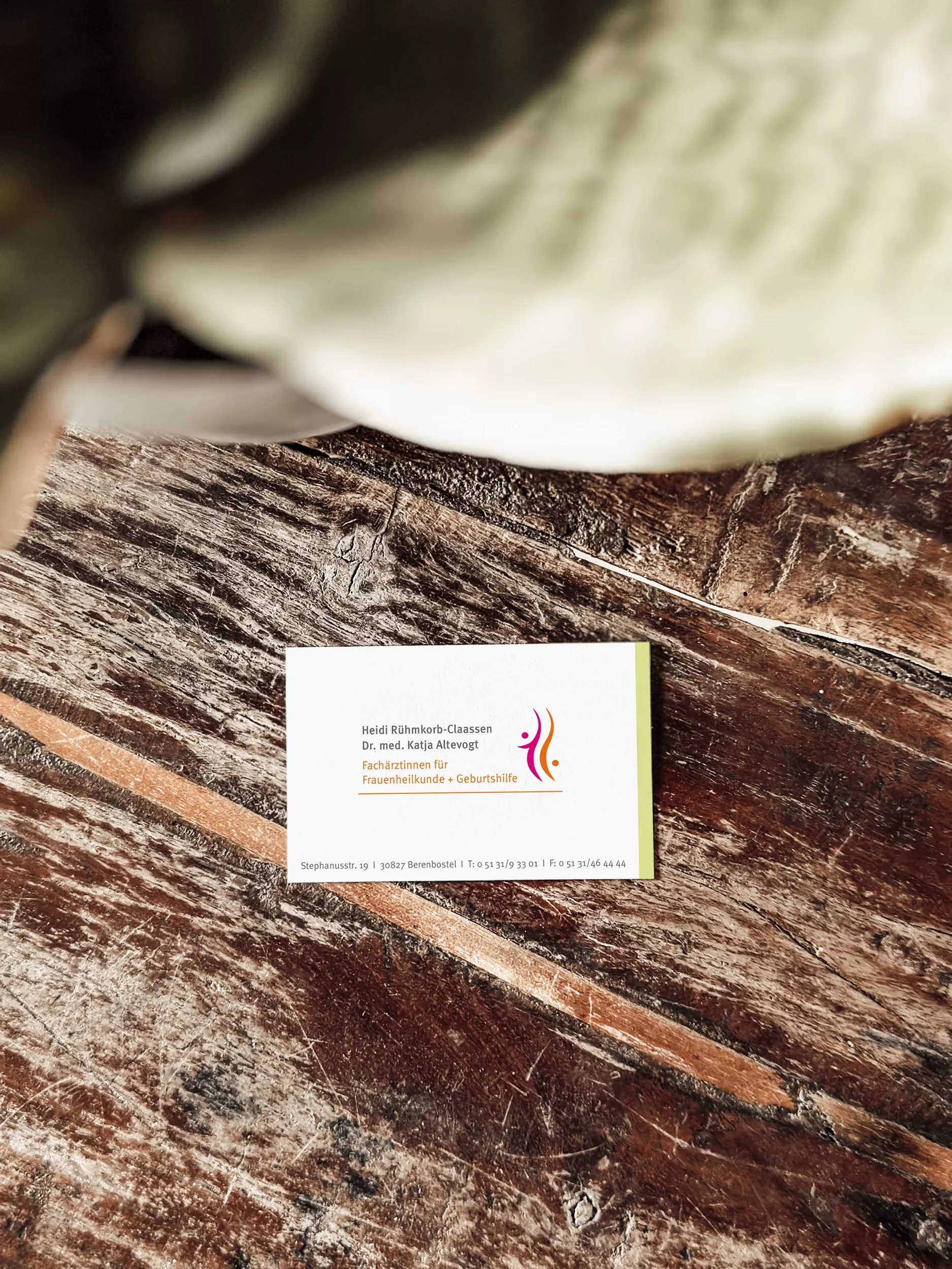 Eine Visitenkarte der Frauenarztpraxis Heidi Rühmkorb-Claassen und Dr. med. Katja Altevogt liegt auf einem rustikalen Holztisch. Die Karte zeigt das Praxislogo mit zwei stilisierten Figuren in Magenta und Orange sowie die Kontaktdaten der Praxis. Das Design wirkt freundlich, modern und professionell.