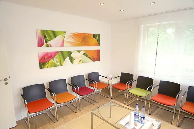 Warteraum der Frauenarztpraxis Dr. Altevogt und Dr. Rühmkorb-Claassen mit modernen Stühlen in warmen Farben (Orange, Rot, Grün) und einer Glasablage mit Getränken. An der weißen Wand hängen zwei farbenfrohe Bilder mit floralen Motiven, die eine freundliche und einladende Atmosphäre schaffen.