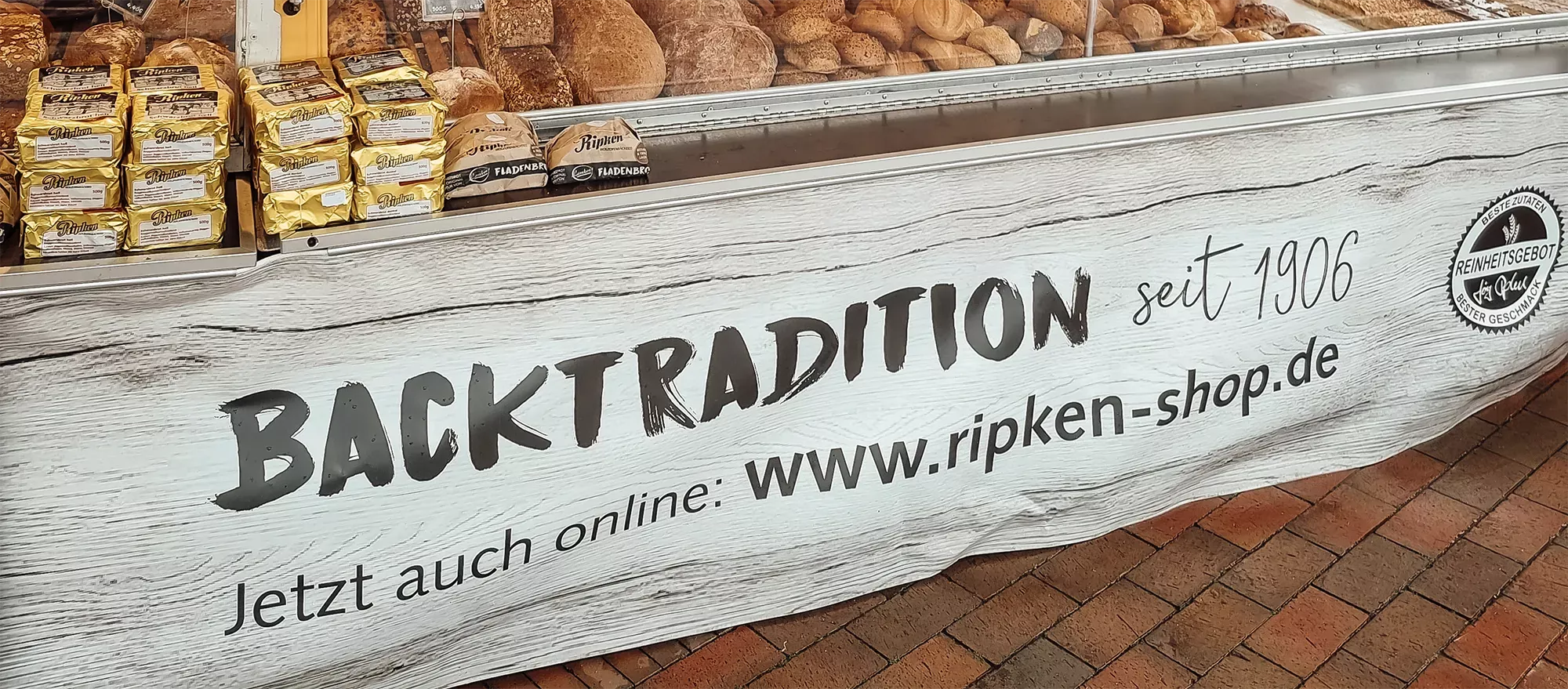 An einem Wochenmarktstand hängt ein Banner, das auf den Onlineshop der Bäckerei verweist.