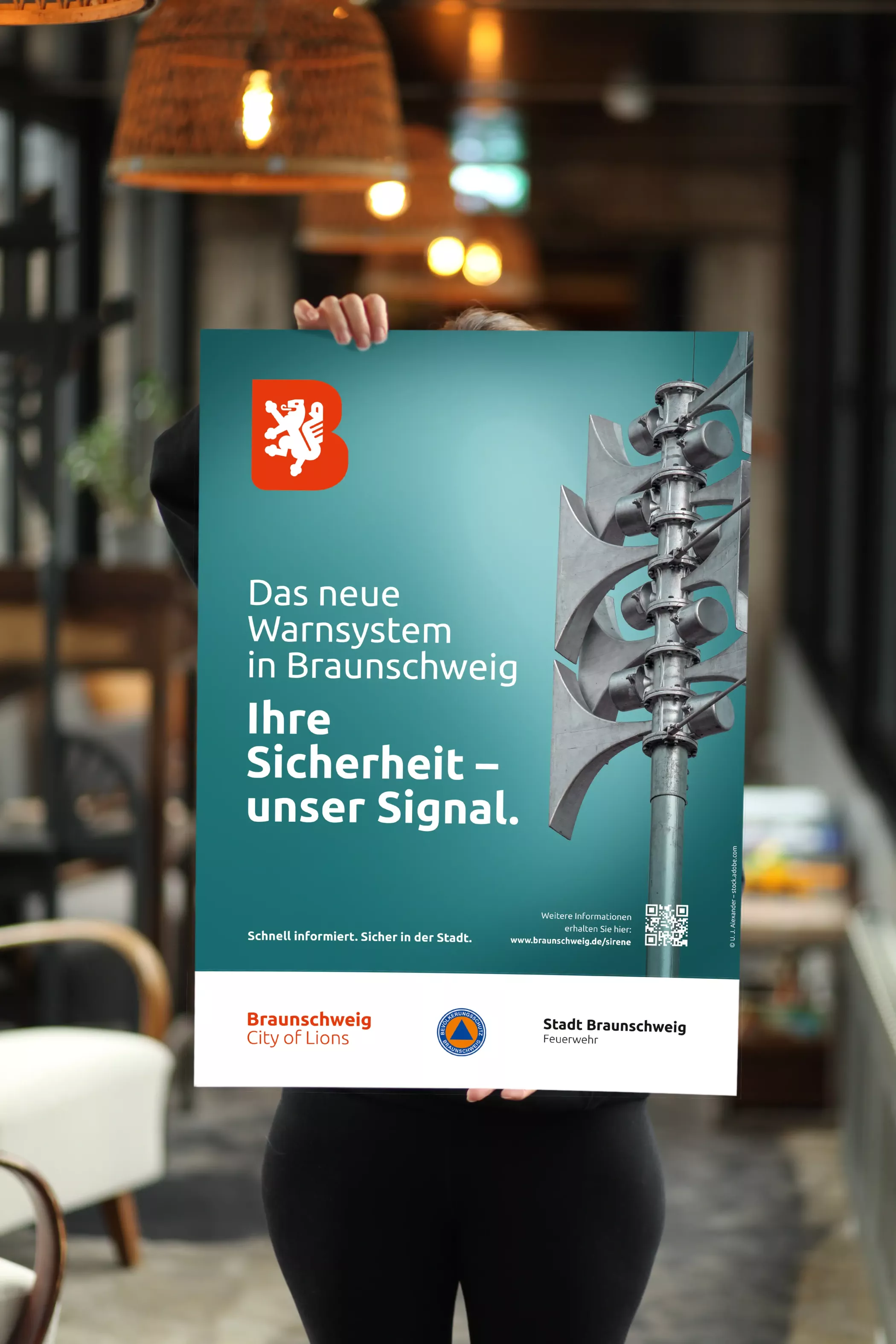 Person hält ein Plakat zur Einführung des neuen Sirenenwarnsystems in Braunschweig mit dem Slogan „Ihre Sicherheit – unser Signal“.