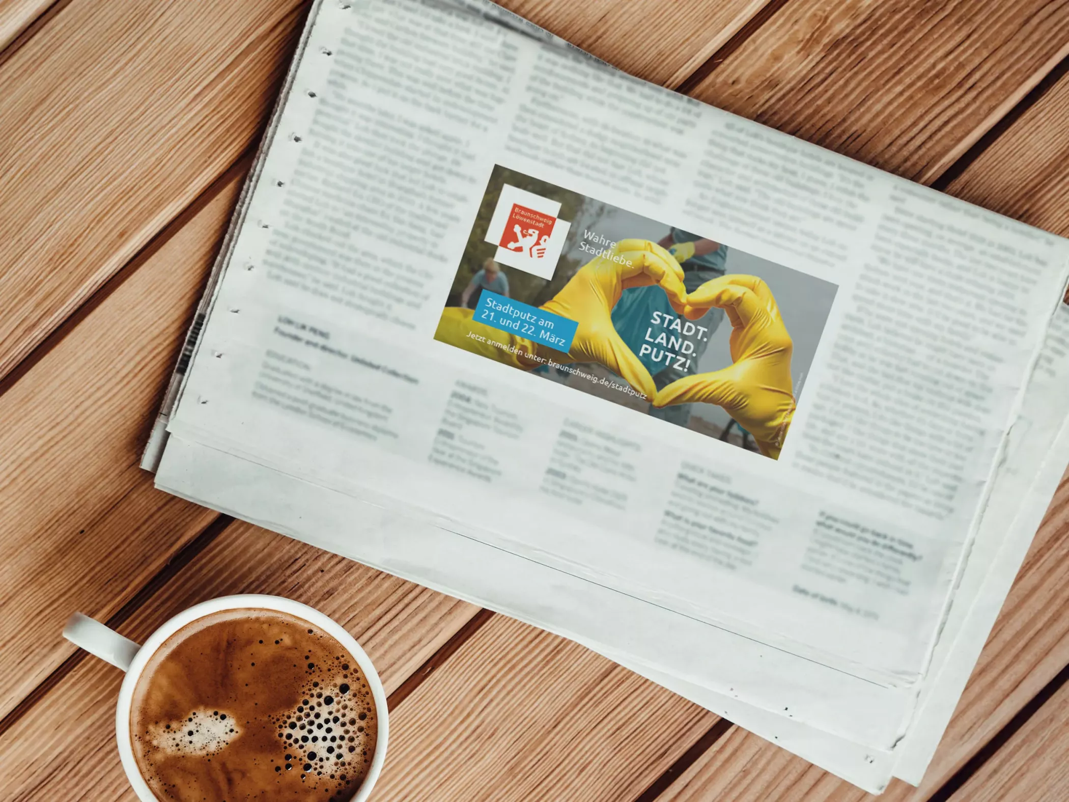 Stadtputz-Anzeige der Stadt Braunschweig in einer Zeitung. Das Motiv zeigt zwei Hände in gelben Handschuhen, die ein Herz formen, daneben der Schriftzug „STADT. LAND. PUTZ.“ sowie Informationen zum Stadtputz am 21. und 22. März. Im Vordergrund steht eine Tasse Kaffee auf einem Holztisch. Die Anzeige wirkt freundlich und aktivierend.