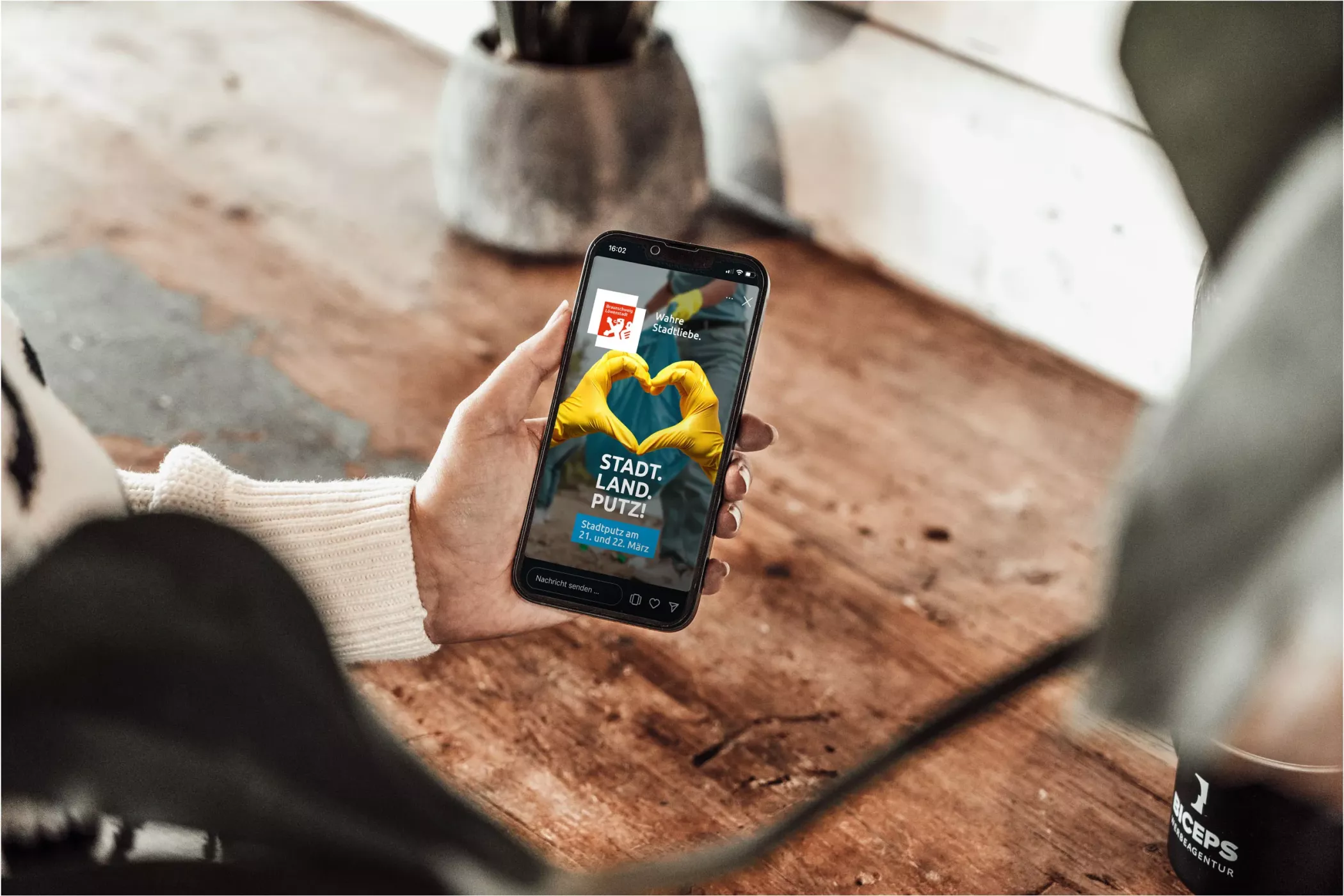 Stadtputz Braunschweig Anzeige auf einem Smartphone-Display. Das Motiv zeigt zwei Hände in gelben Handschuhen, die ein Herz formen, mit dem Schriftzug „STADT. LAND. PUTZ.“ sowie Informationen zum Stadtputz am 21. und 22. März. Im Hintergrund eine Hand, die das Handy hält, auf einem Holztisch mit Pflanze und Becher.