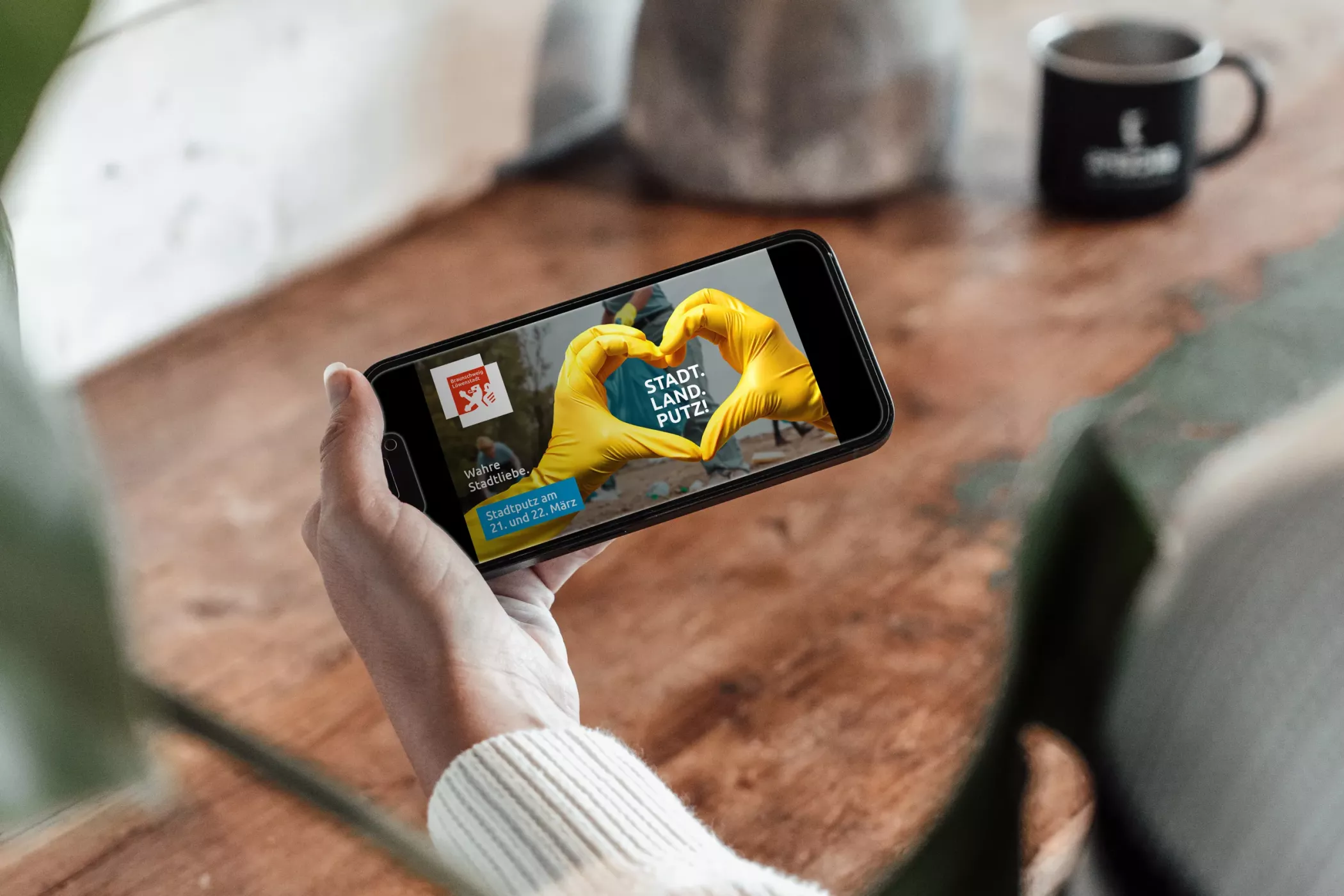 Digitale Stadtputz-Anzeige der Stadt Braunschweig auf einem Smartphone im Querformat. Das Motiv zeigt zwei Hände in gelben Handschuhen, die ein Herz formen, dazu den Schriftzug „STADT. LAND. PUTZ.“ und Informationen zum Stadtputz am 21. und 22. März. Das Handy wird von einer Person über einem Holztisch gehalten, im Hintergrund sind Pflanzen und eine Tasse zu sehen.