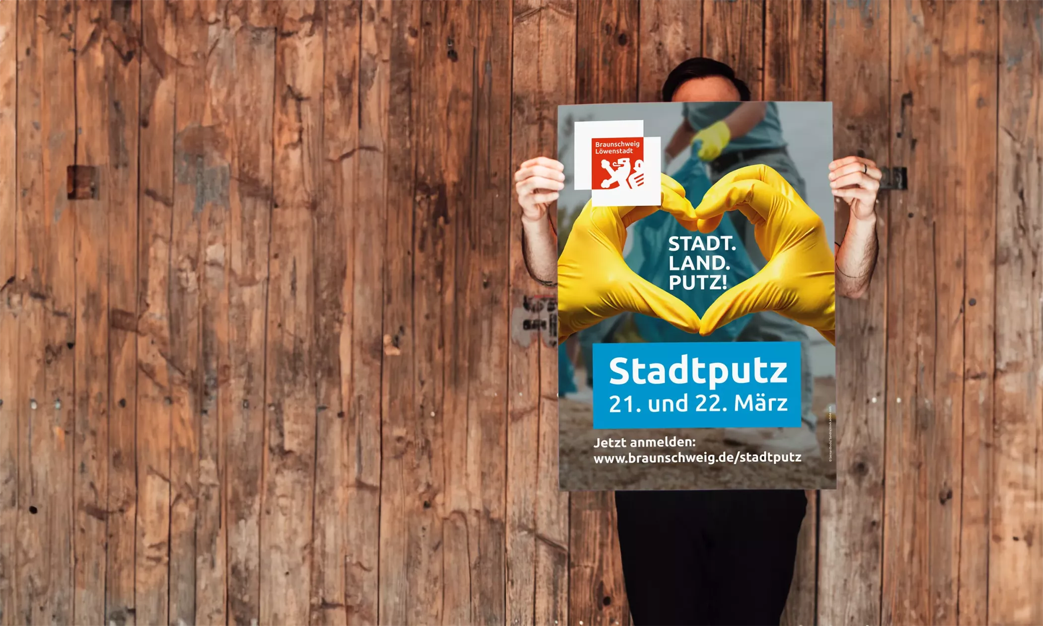 Eine Person hält ein Stadtputz-Plakat der Stadt Braunschweig vor einer Holzwand. Auf dem Plakat formen zwei Hände in gelben Handschuhen ein Herz. Der Slogan „STADT. LAND. PUTZ.“ sowie Informationen zum Stadtputz am 21. und 22. März und der Link zur Anmeldung sind gut sichtbar. Das Motiv wirkt freundlich, motivierend und aktivierend.