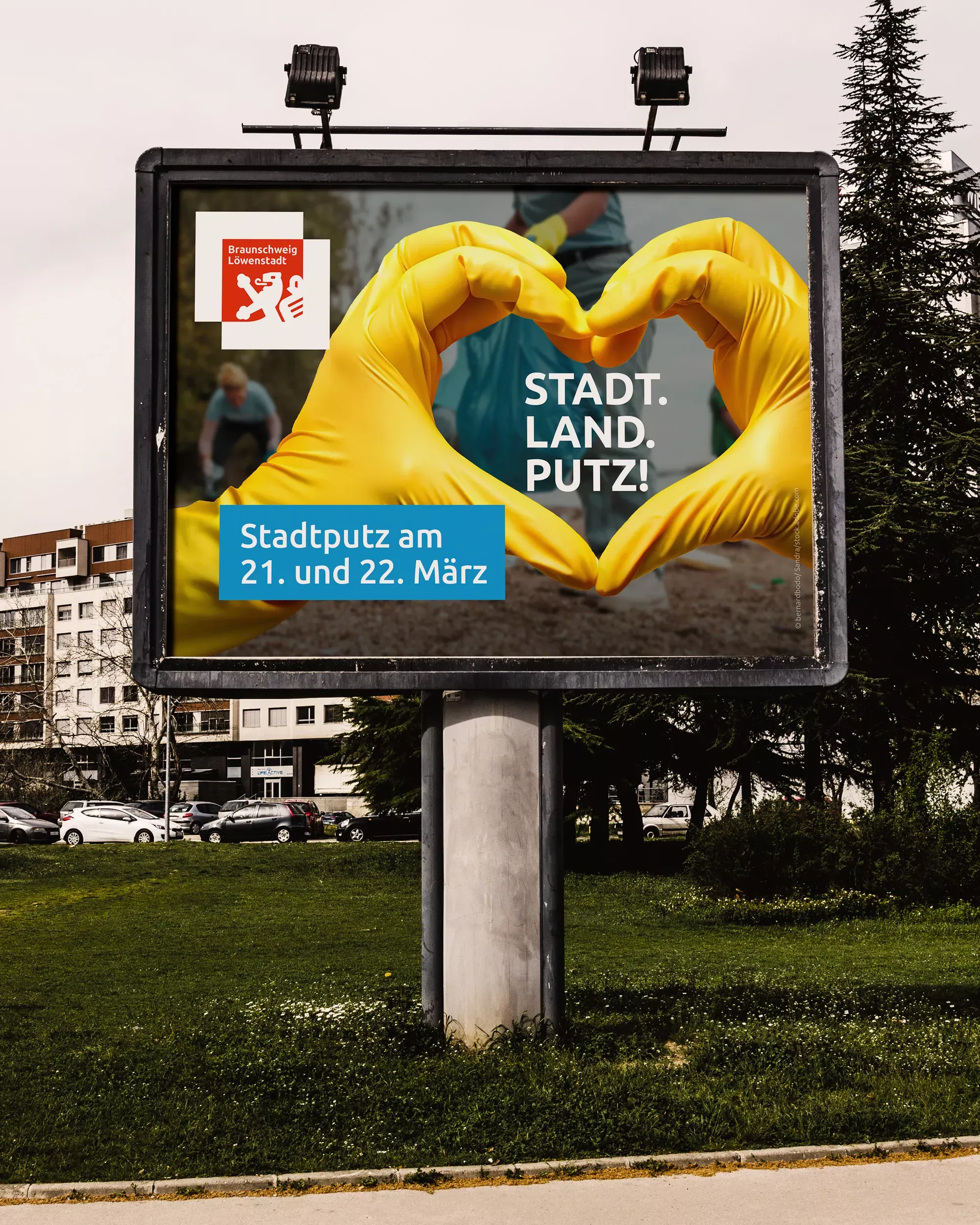 Großflächenplakat für den Stadtputz Braunschweig auf einer Werbetafel im Freien. Das Motiv zeigt zwei Hände in gelben Handschuhen, die ein Herz formen, dazu den Schriftzug „STADT. LAND. PUTZ.“ und den Hinweis auf den Stadtputz am 21. und 22. März. Im Hintergrund sind Wohnhäuser und eine Grünfläche zu sehen.