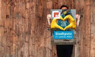 Eine Person hält ein Stadtputz-Plakat der Stadt Braunschweig vor einer Holzwand. Auf dem Plakat formen zwei Hände in gelben Handschuhen ein Herz. Der Slogan „STADT. LAND. PUTZ.“ sowie Informationen zum Stadtputz am 21. und 22. März und der Link zur Anmeldung sind gut sichtbar. Das Motiv wirkt freundlich, motivierend und aktivierend.