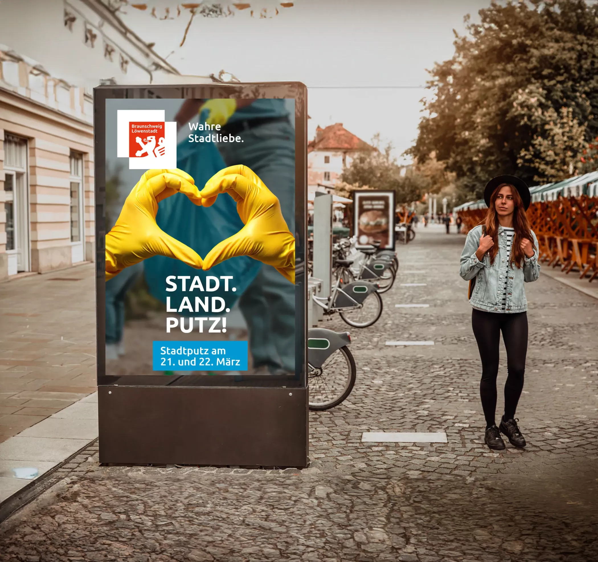 Digitale Stadtputz-Anzeige der Stadt Braunschweig in einer Fußgängerzone. Auf dem Motiv formen zwei Hände in gelben Handschuhen ein Herz, dazu der Schriftzug „STADT. LAND. PUTZ.“ sowie der Hinweis auf den Stadtputz am 21. und 22. März. Im Vordergrund läuft eine junge Frau vorbei, im Hintergrund stehen Fahrräder und weitere Werbedisplays.