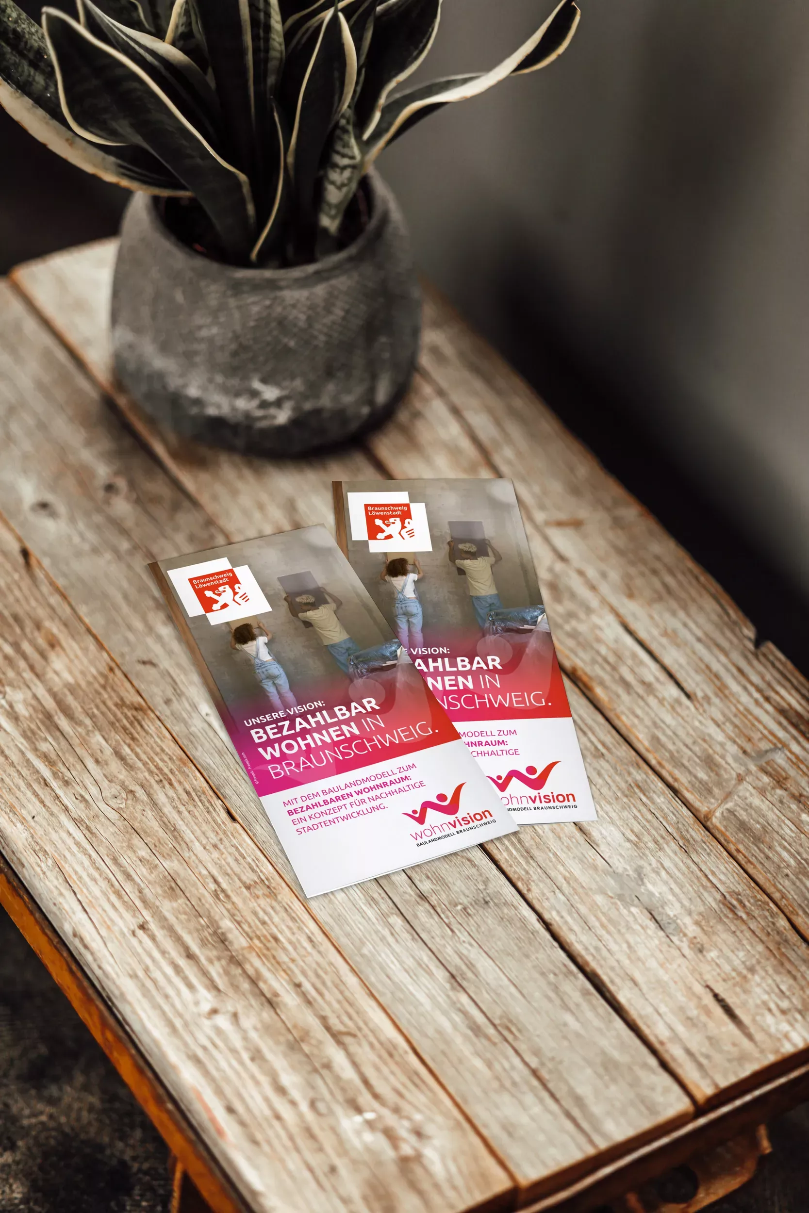 Zwei Wohnvision-Faltblätter liegen auf einem rustikalen Holztisch. Auf dem Titel steht in großen Lettern „Bezahlbar wohnen in Braunschweig“. Im Hintergrund ist eine Frau mit Umzugskarton zu sehen, vorne das Wohnvision-Logo und der Löwenstadt-Schriftzug.