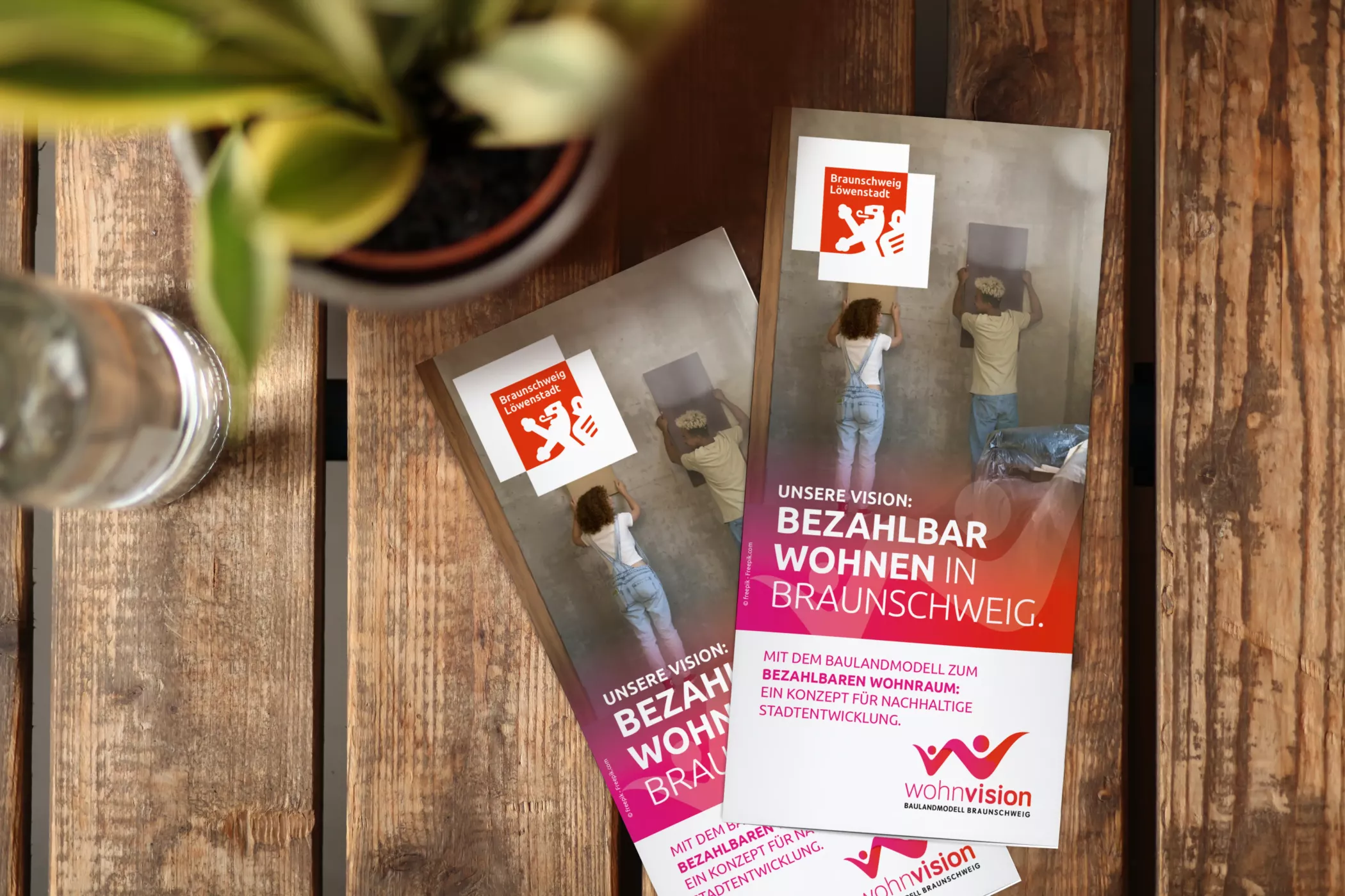 Zwei Exemplare der wohnvision-Broschüre liegen auf einem rustikalen Holztisch, daneben ein Glas Wasser und eine Pflanze. Die Titel zeigen zwei umziehende Menschen mit Kartons sowie die Headline: „Unsere Vision: Bezahlbar wohnen in Braunschweig.“