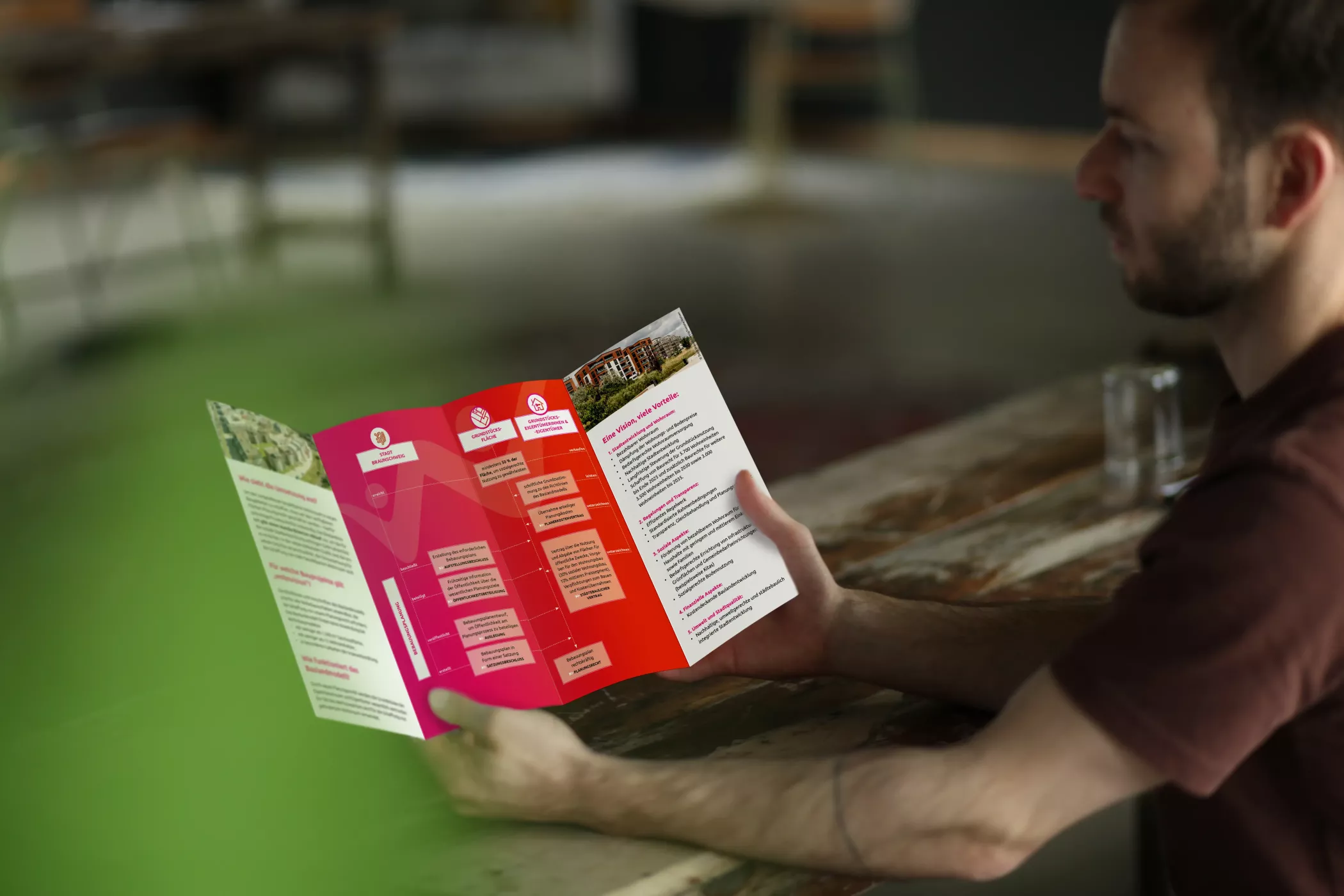 Ein Mann hält den geöffneten wohnvision-Flyer in der Hand. Sichtbar sind die Innenseiten mit anschaulichen Grafiken und Textblöcken, die den Ablauf des neuen Baulandmodells in Braunschweig erklären. Die Rückseite greift das Farbkonzept der Kampagne in Rot- und Pinktönen auf.