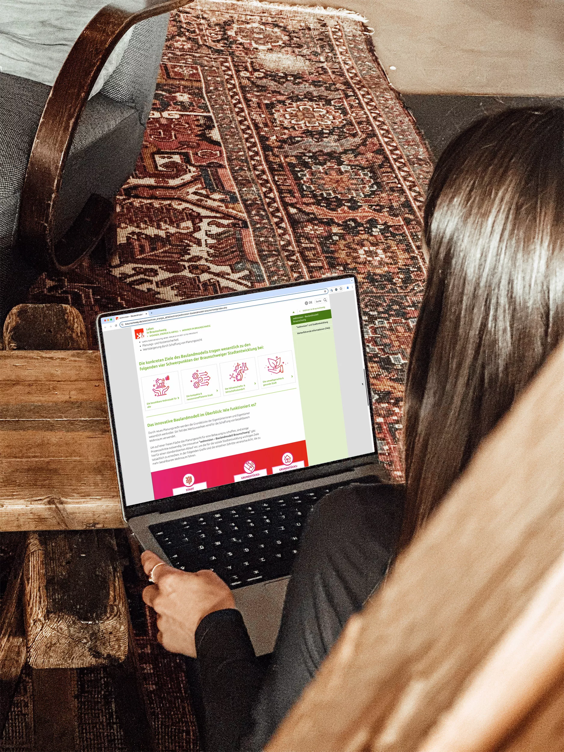 Eine Frau sitzt auf einem Teppichboden und arbeitet an einem Laptop. Auf dem Bildschirm ist die Website der Stadt Braunschweig geöffnet, auf der das Wohnvision-Baulandmodell erklärt wird.