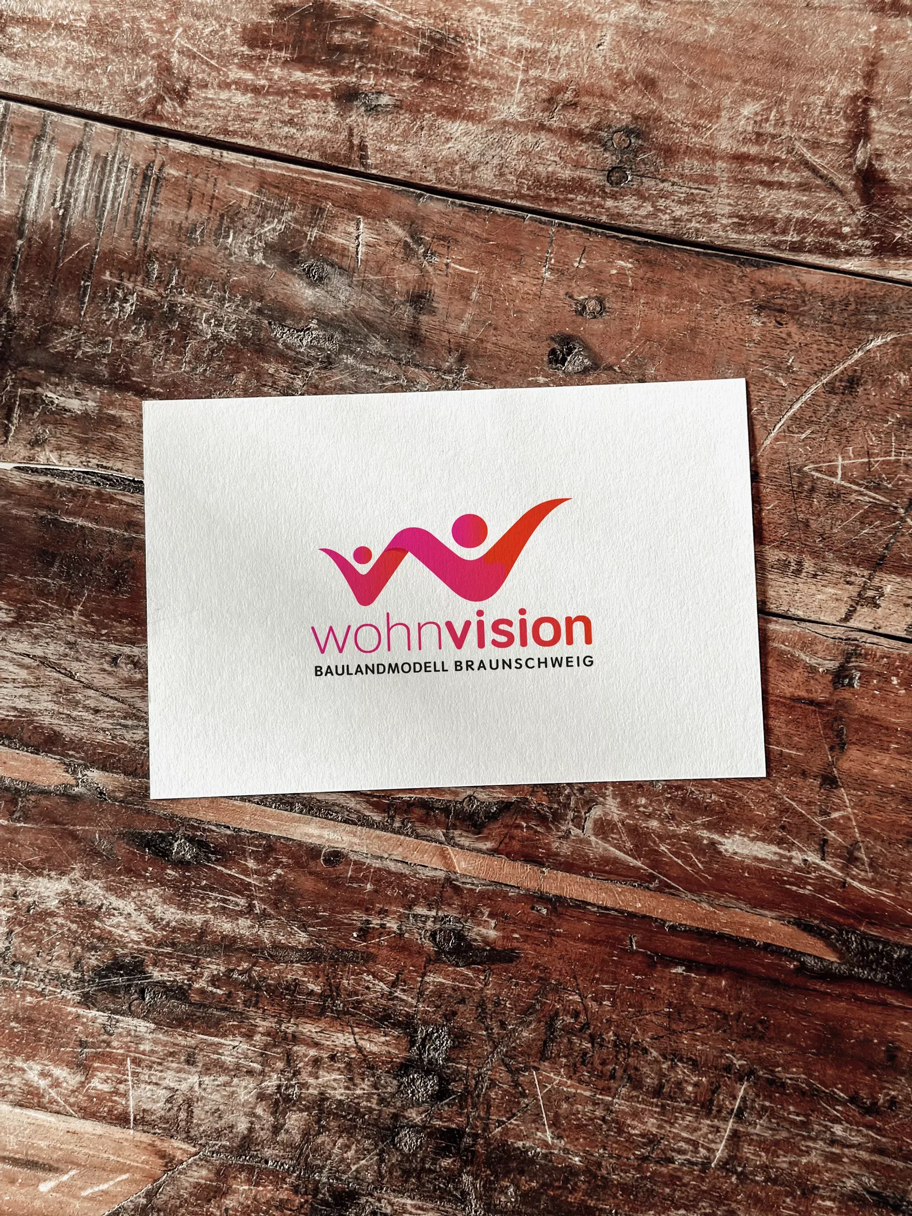 Karte mit dem Logo der Wohnvision Braunschweig auf einem Holztisch. Das farbenfrohe Logo zeigt zwei stilisierte Figuren in Rot- und Violetttönen, die sich dynamisch zu einem abstrahierten Haus mit zentraler Kreisform verbinden. Darunter steht der Schriftzug „wohnvision – Baulandmodell Braunschweig“.