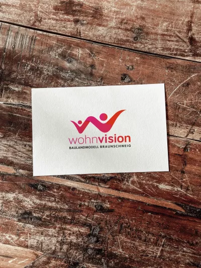 Karte mit dem Logo der Wohnvision Braunschweig auf einem Holztisch. Das farbenfrohe Logo zeigt zwei stilisierte Figuren in Rot- und Violetttönen, die sich dynamisch zu einem abstrahierten Haus mit zentraler Kreisform verbinden. Darunter steht der Schriftzug „wohnvision – Baulandmodell Braunschweig“.