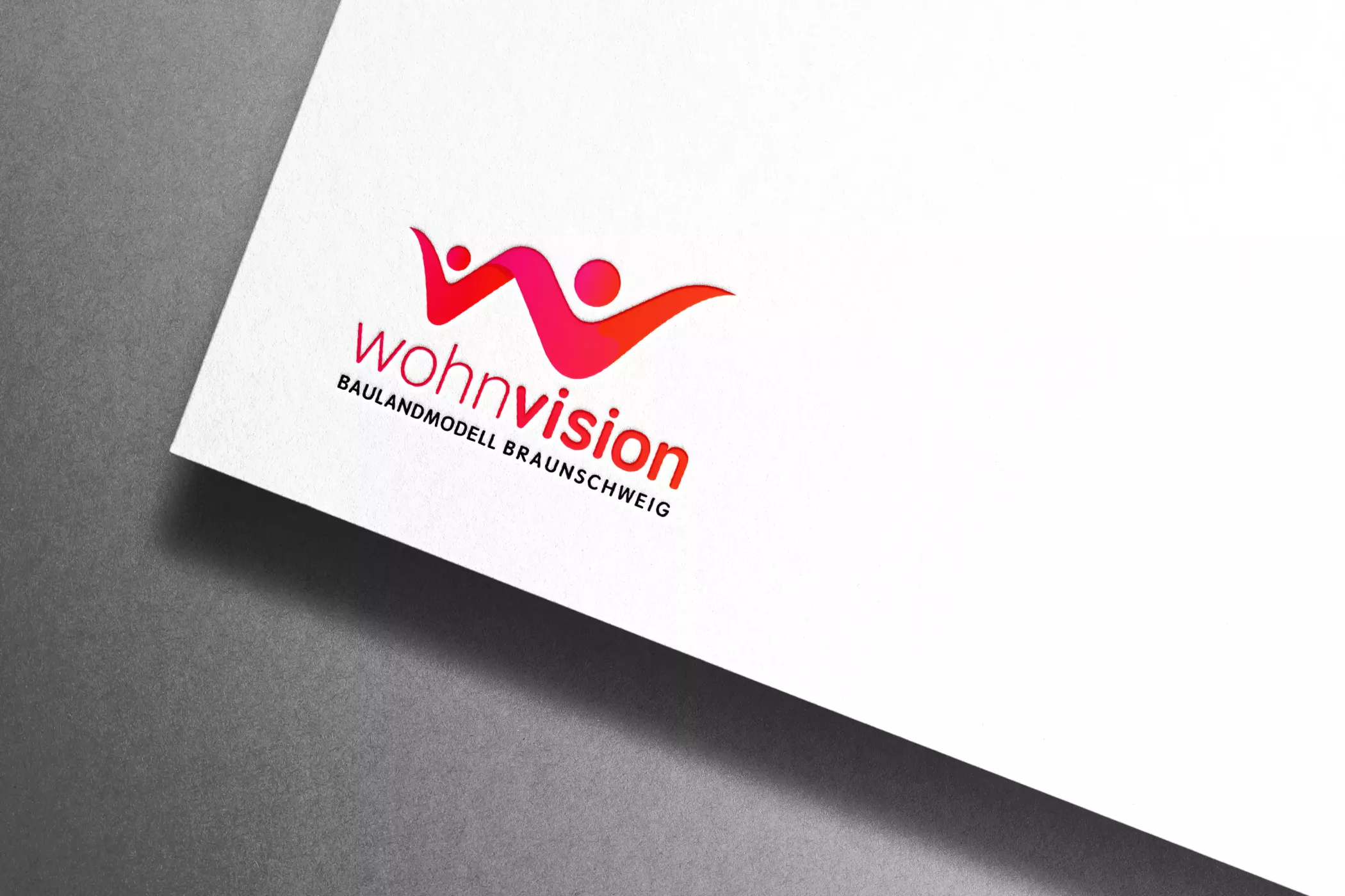 Detailansicht des Wohnvision-Logos auf weißem Papier vor grauem Hintergrund. Das Logo zeigt zwei abstrahierte Figuren in kräftigem Pink und Rot, die zusammen ein Dach formen. Der Schriftzug „wohnvision – Baulandmodell Braunschweig“ steht in modernem, klaren Design darunter.
